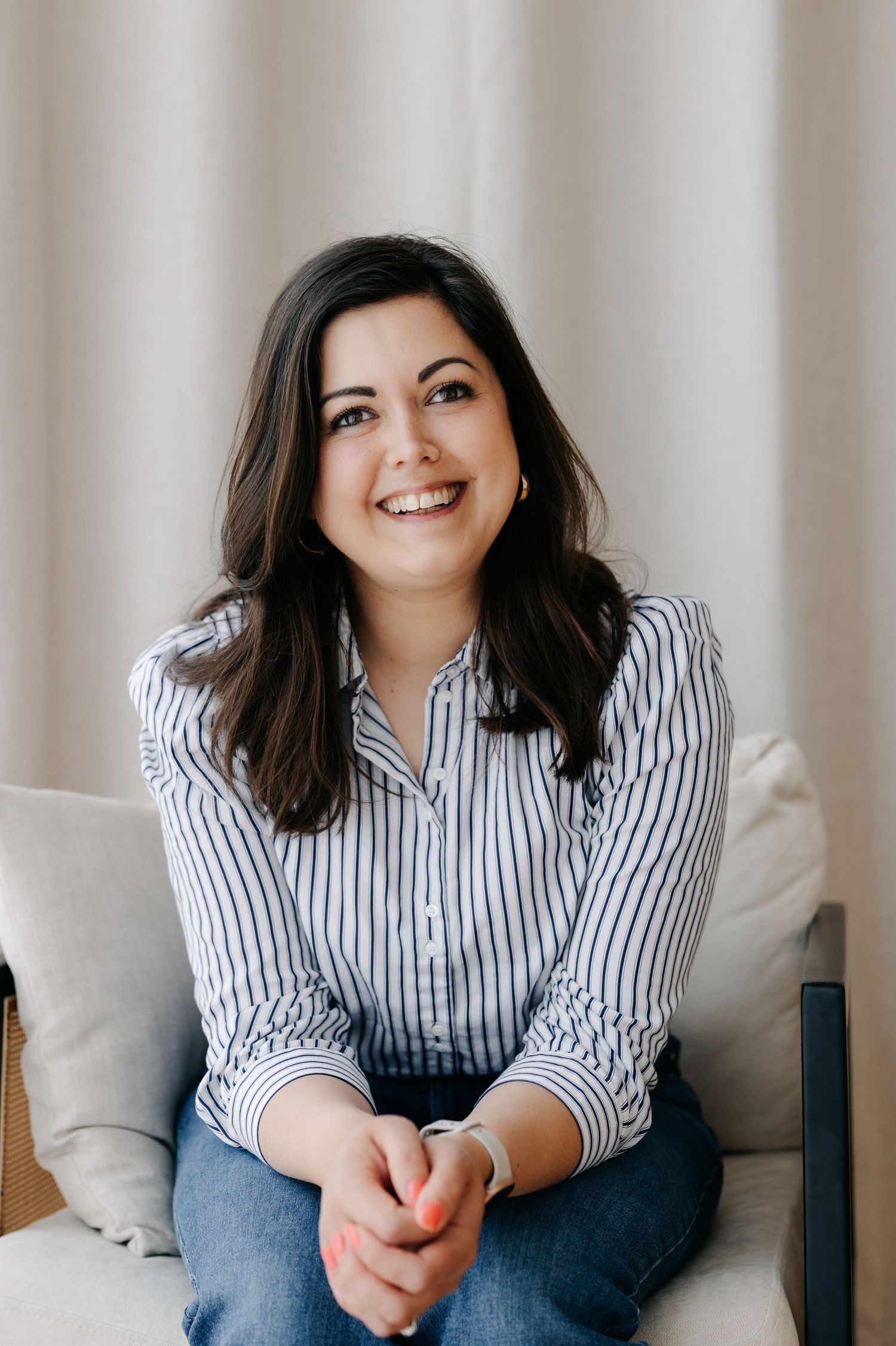 Smiling brunette woman in striped shirt sitting relaxed on a couch with hands clasped, natural light background.