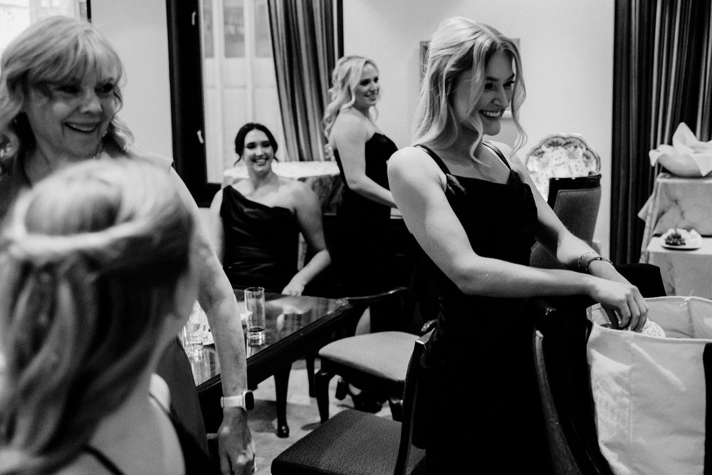 Bridesmaids in black dresses laugh and celebrate together in a candid black and white wedding preparation photo.