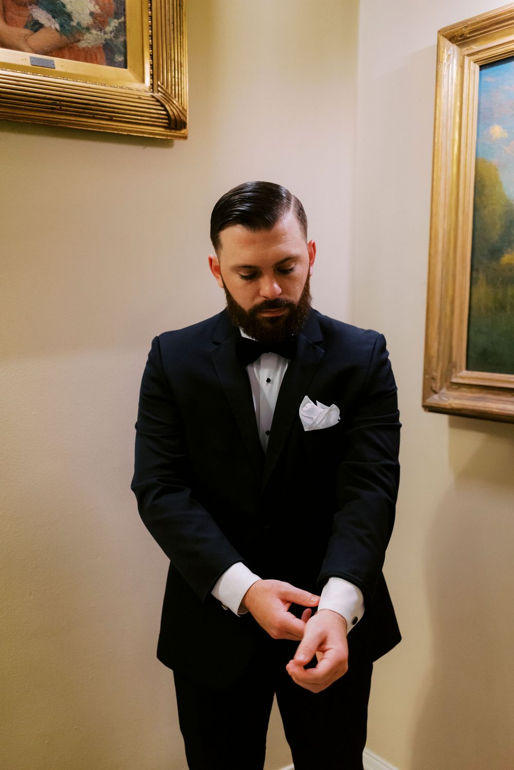 Bearded groom in black tuxedo with bow tie and white pocket square adjusting cufflinks between artistic paintings before wedding ceremony.