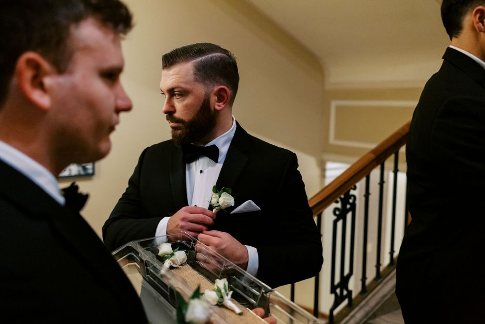 Layered, photojournalistic photo of groomsman in black tuxedo pinning boutonniere near staircase before wedding ceremony.