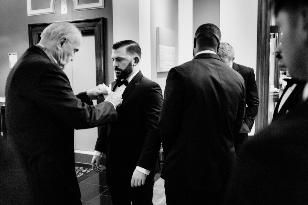 Monochrome photo of groomsmen in black tuxedos help each other with boutonnieres before a wedding ceremony.