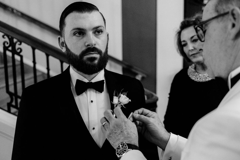 Black and white filmic photo of a bearded groom in a tuxedo having his boutonniere pinned on before his Chicago Cathedral wedding.