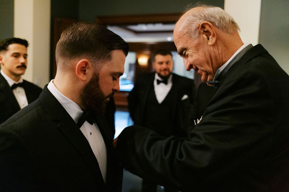 Elderly man carefully pins a boutonniere on a groom in a black tuxedo before wedding.