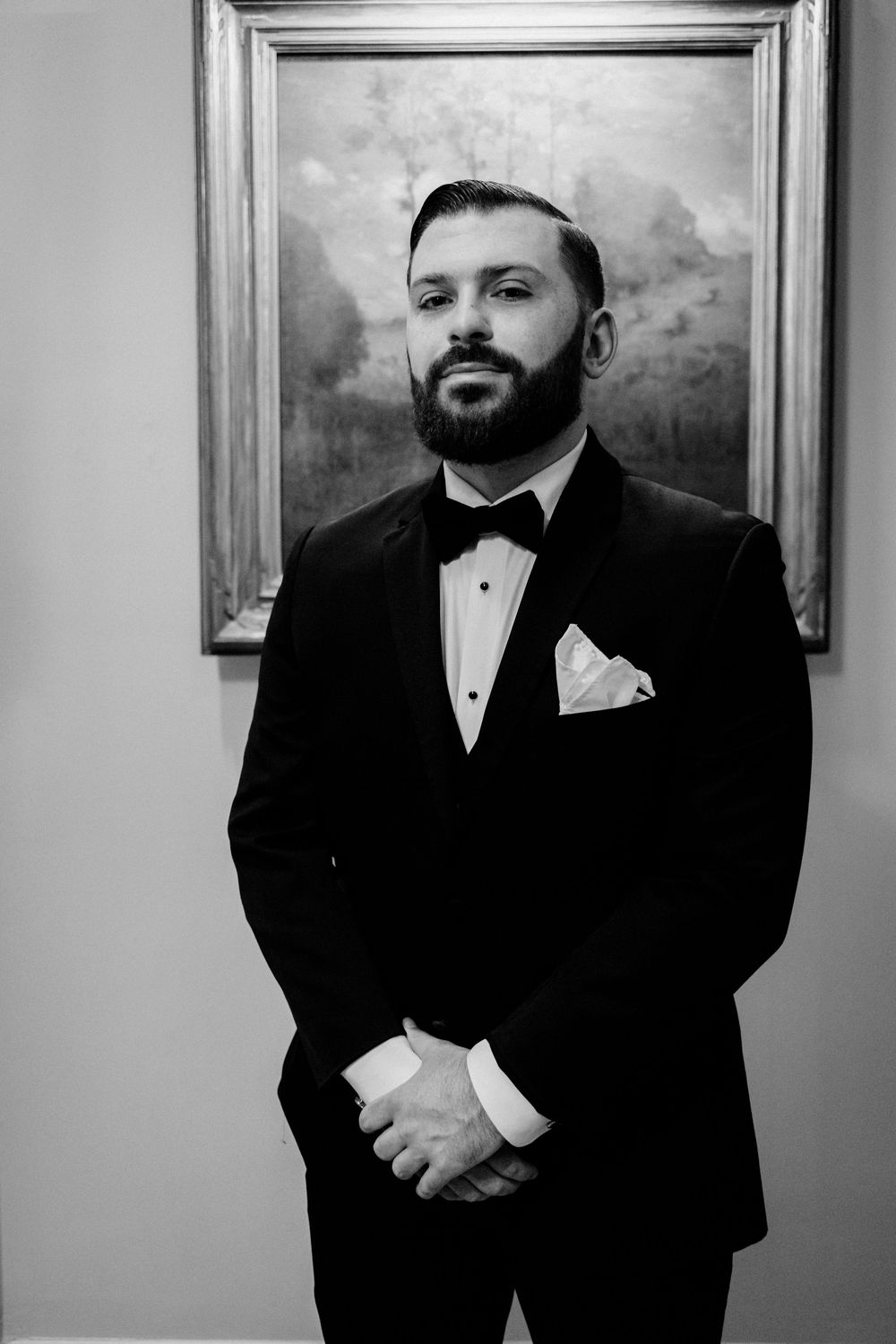 Elegant bearded groom in black tuxedo and bow tie posing confidently in front of painting in black and white portrait image.