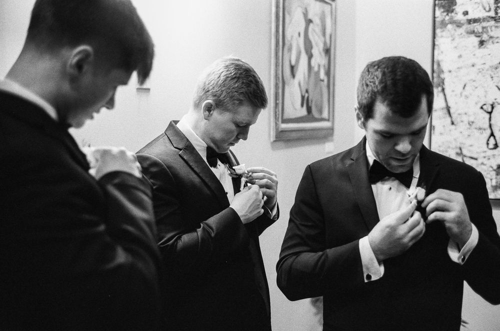 Three groomsmen in black tuxedos adjusting boutonnieres before a wedding ceremony in black and white 35mm film photo.