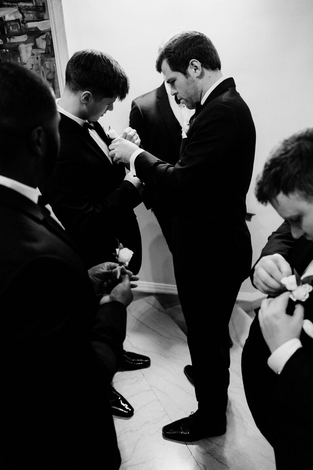 Groomsmen in black tuxedos helping each other pin boutonnieres before a wedding in black and white photo.