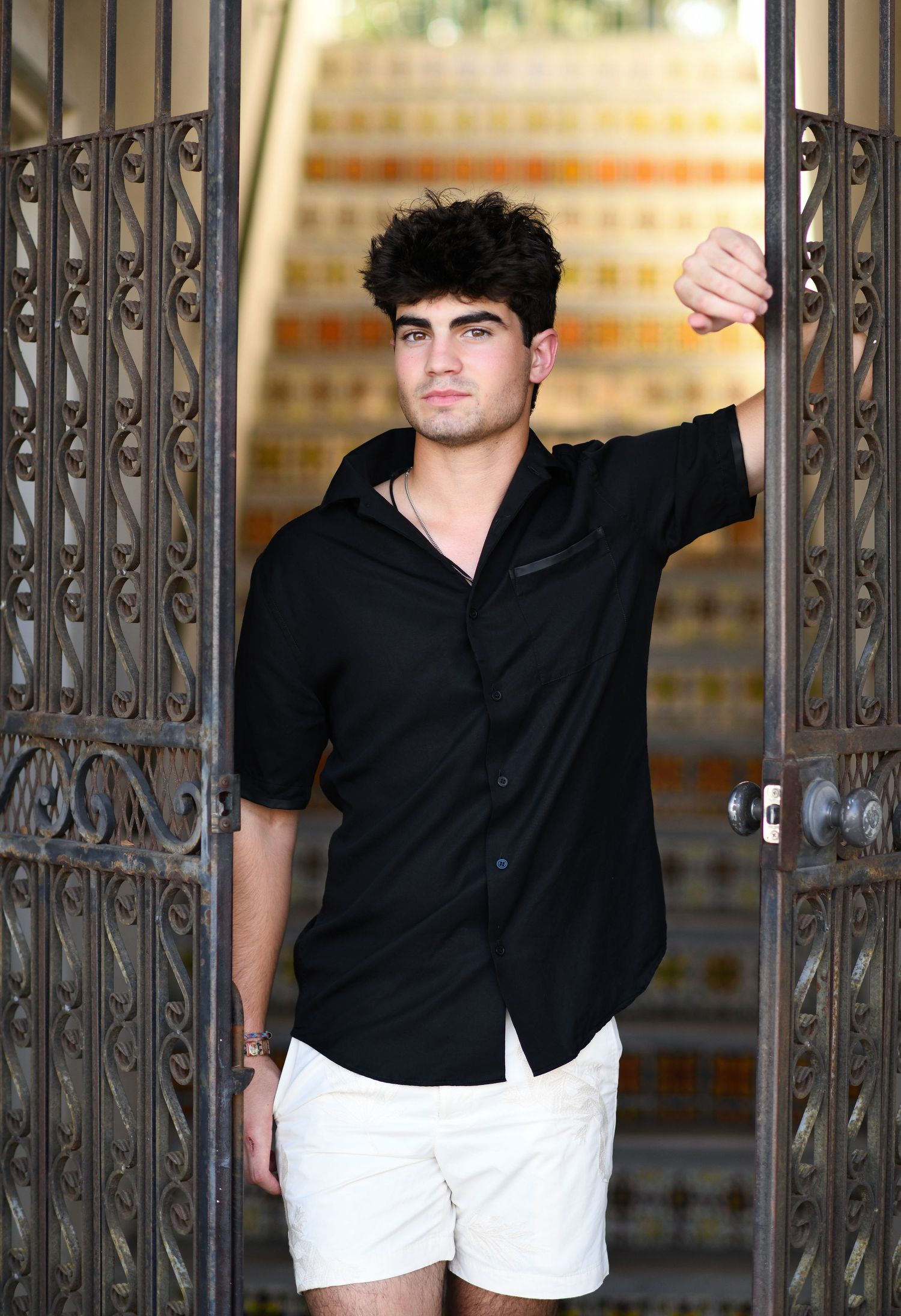 Young man in black shirt and white shorts posing by ornate iron gates.