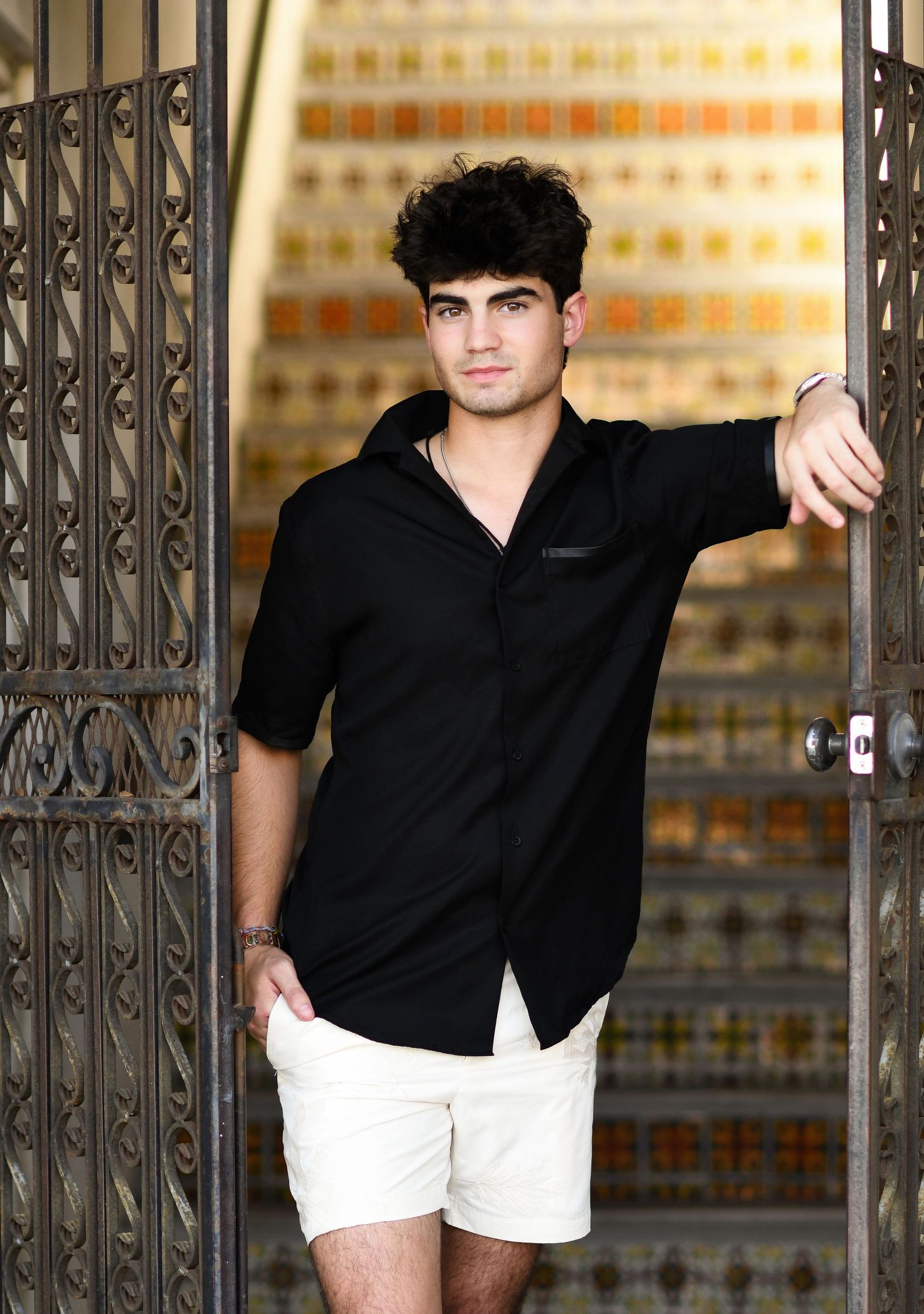 Young man in black shirt and white shorts posing confidently by ornate iron gates with colorful tiled wall backdrop.