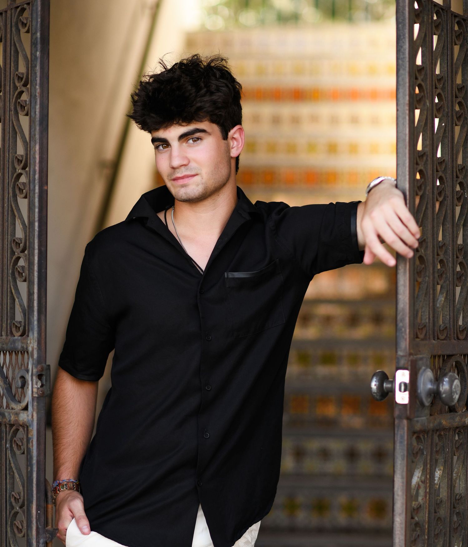 Young man in black shirt leaning against ornate iron doors in a stylish outdoor portrait photo.