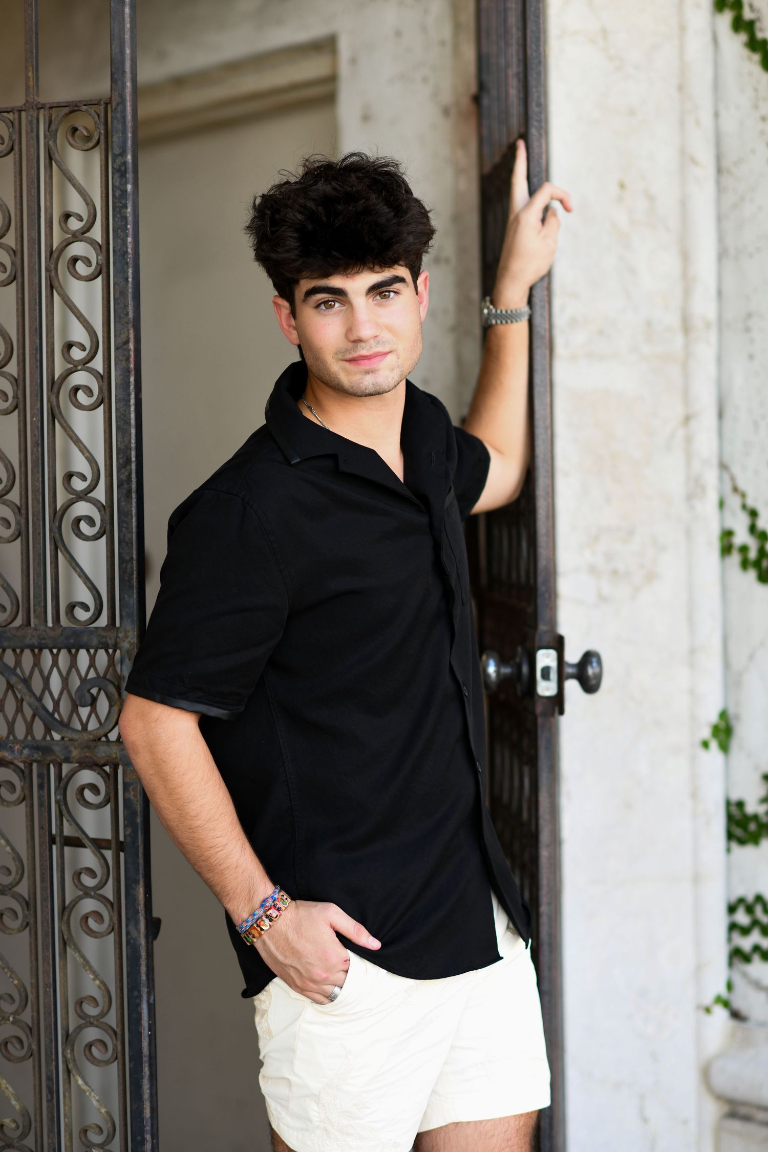 Young man in black shirt and white shorts leaning against ornate iron door in elegant architectural setting.