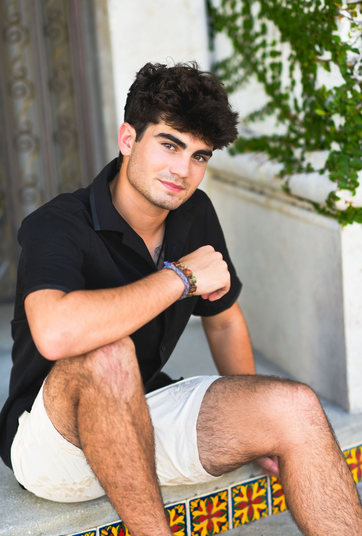 Young man in black shirt and white shorts sitting on decorative tiled steps outdoors.
