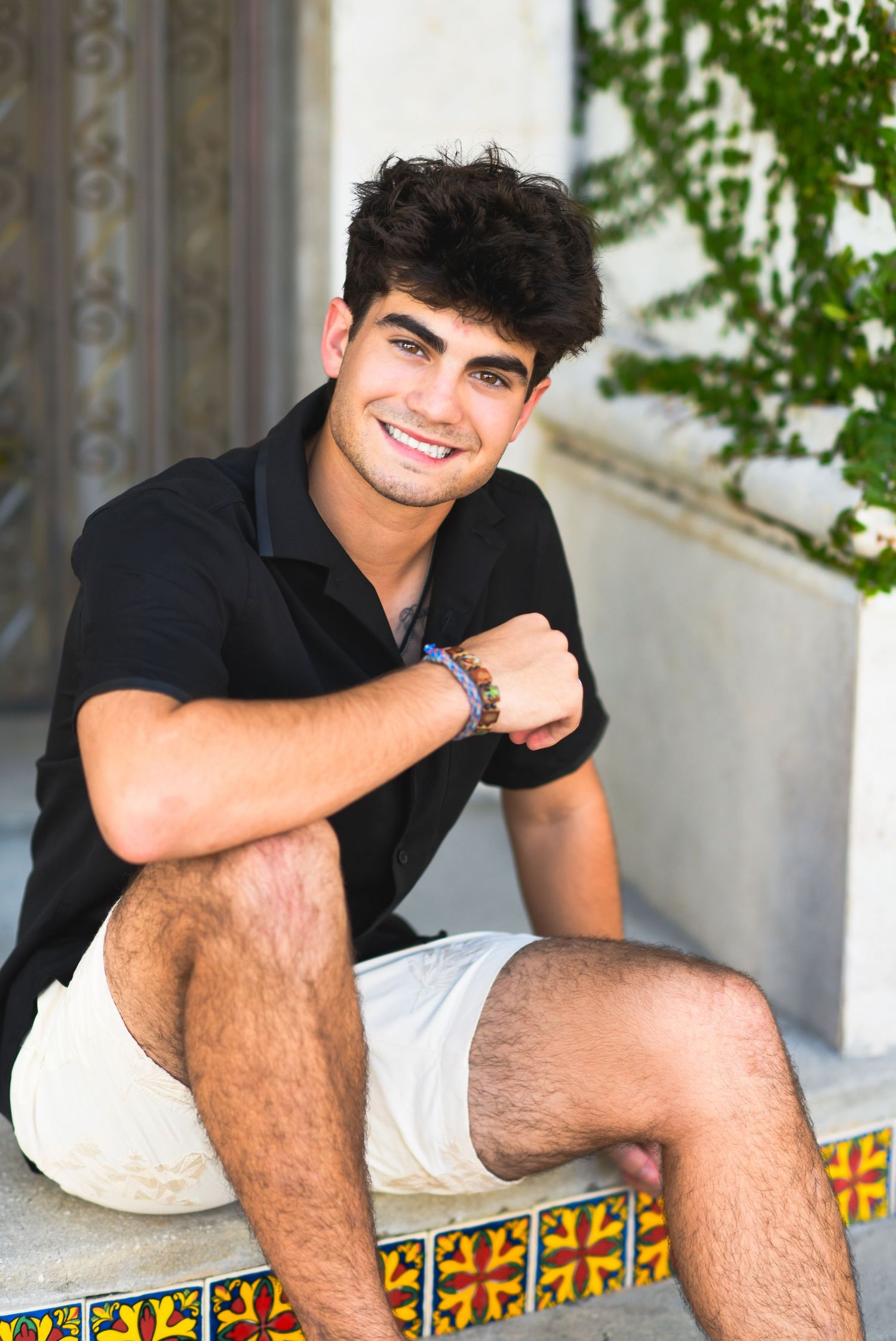 Young man smiling in black shirt and white shorts, sitting on colorful tiled steps outdoors.