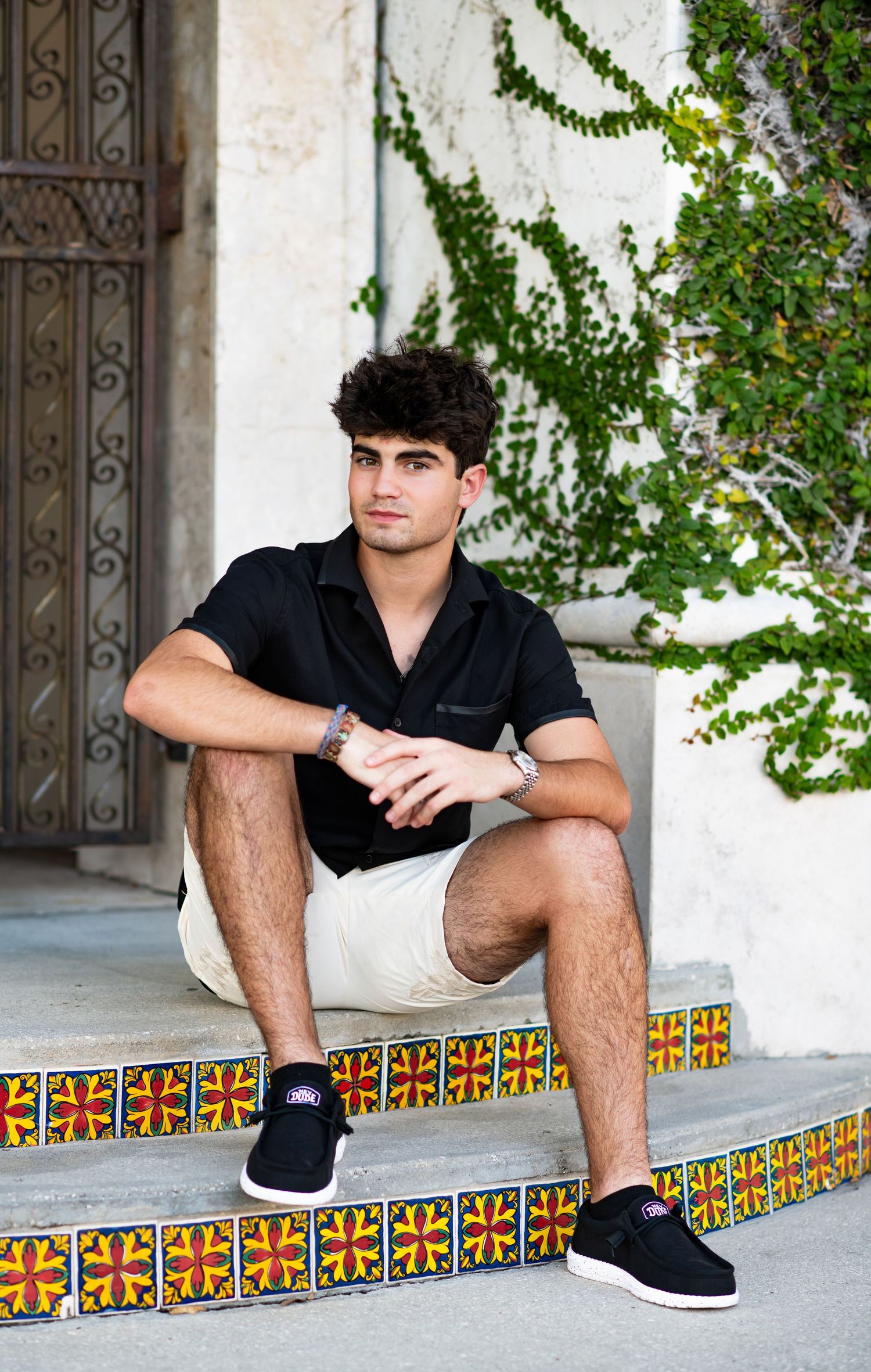 Stylish young man in black polo and khaki shorts sitting on decorative tiled steps outdoors.