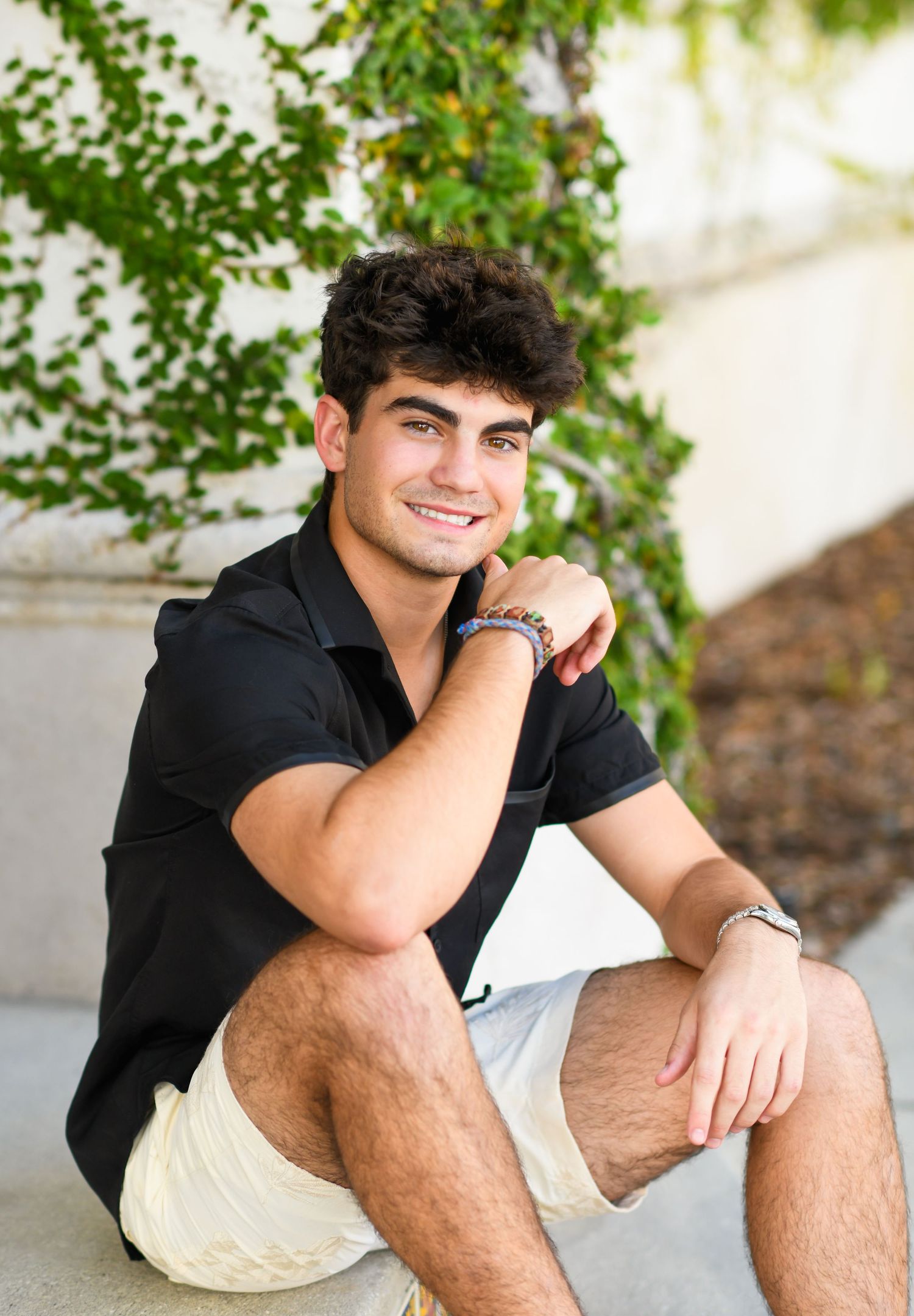 Young man in black polo and white shorts sitting on steps smiling, with ivy wall background.