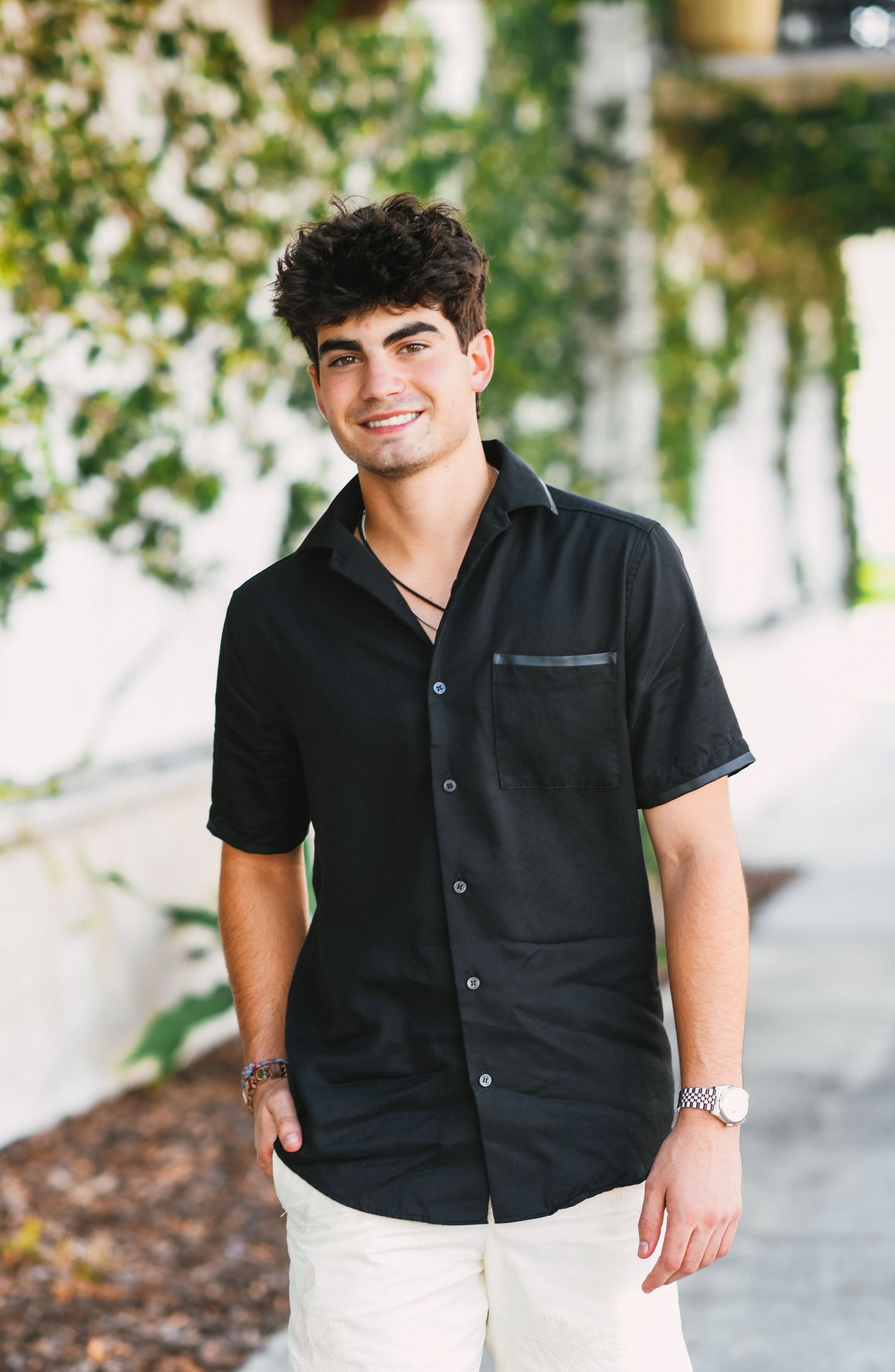 Young man smiling in black short-sleeve button-up shirt and khaki pants outdoors.