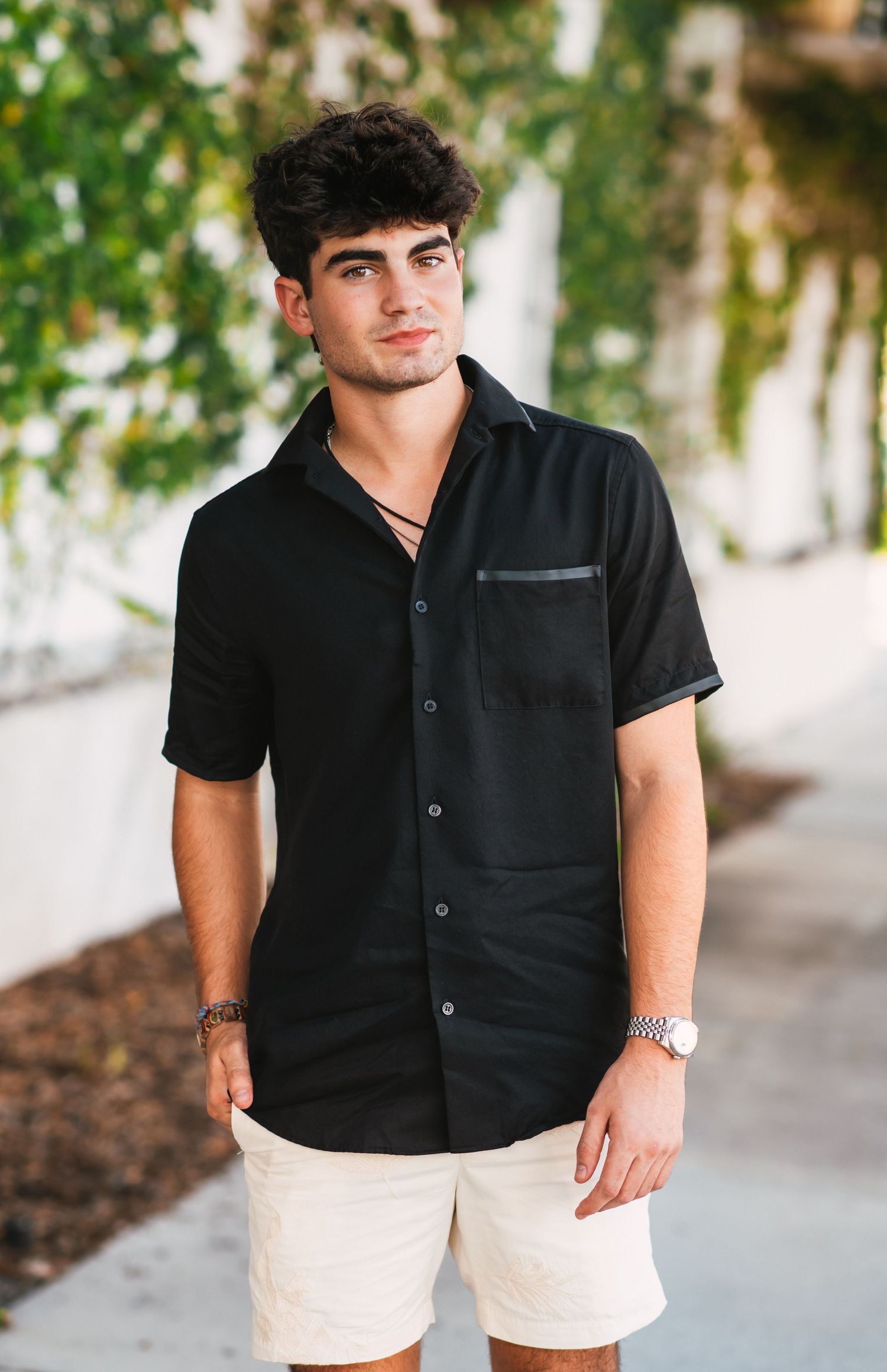 Stylish young man in black button-up shirt and white shorts standing outdoors near greenery.