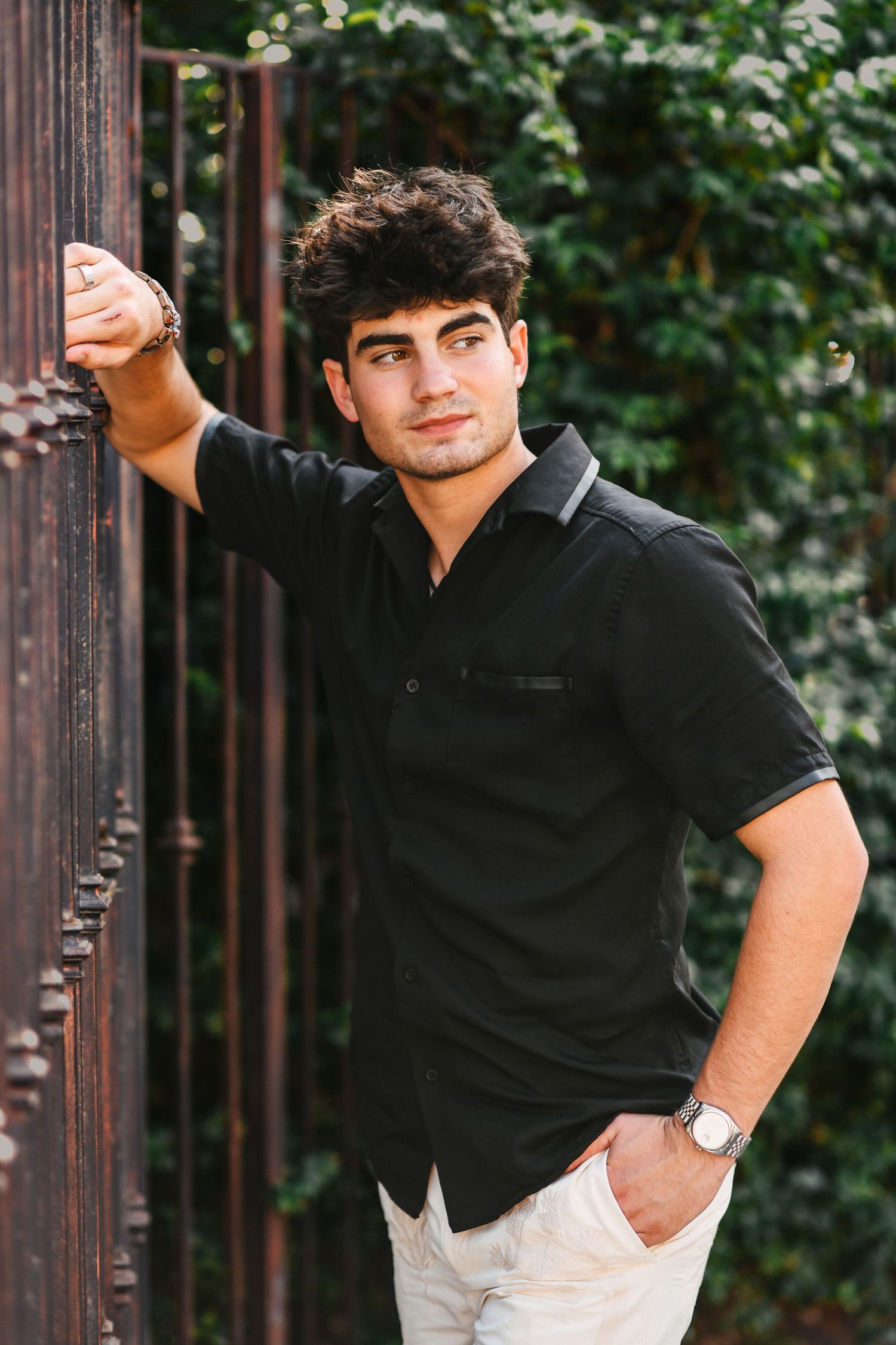 Stylish young man in black shirt leaning on rustic metal fence outdoors.