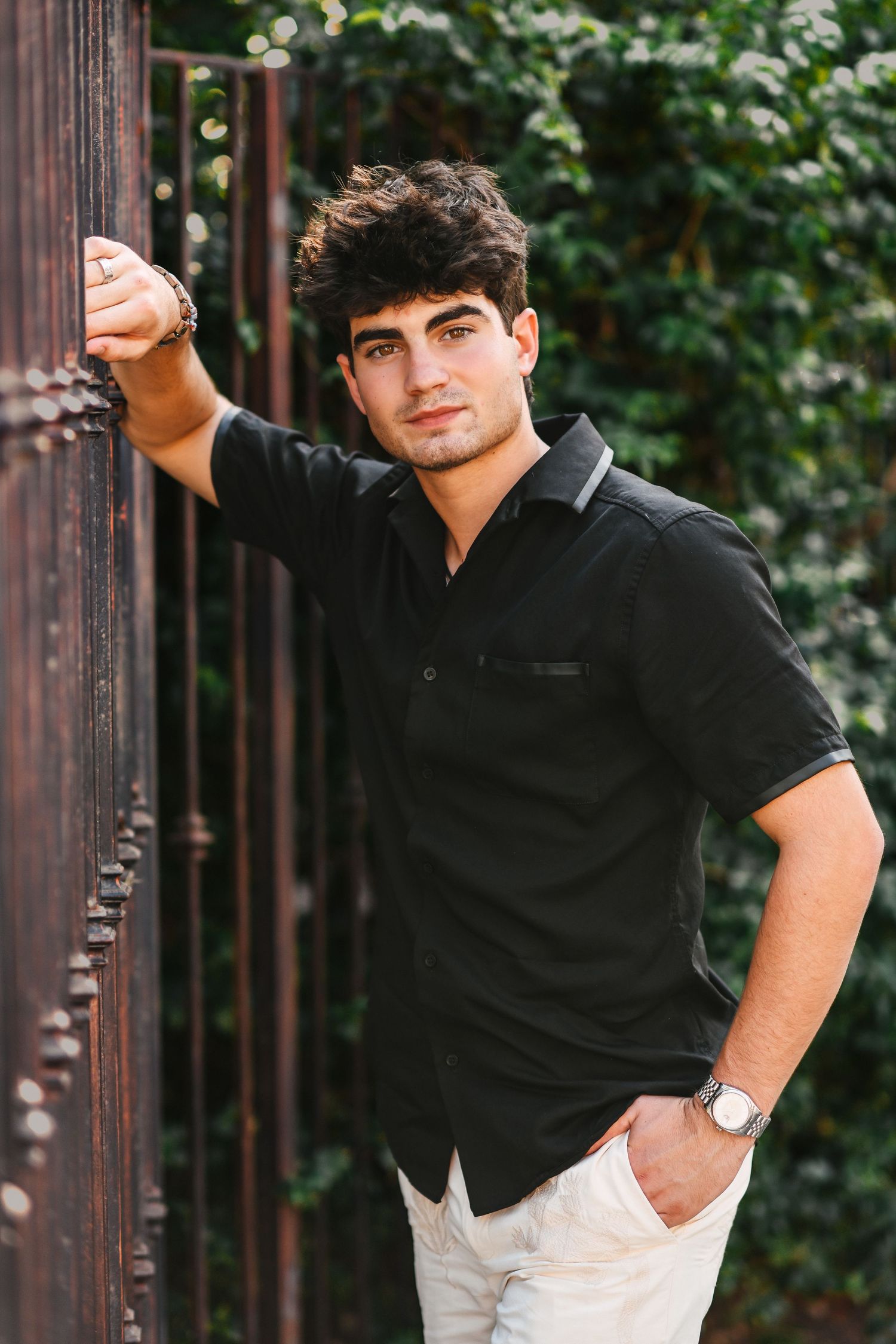 Young man in black shirt leaning against rustic metal fence with green foliage background.