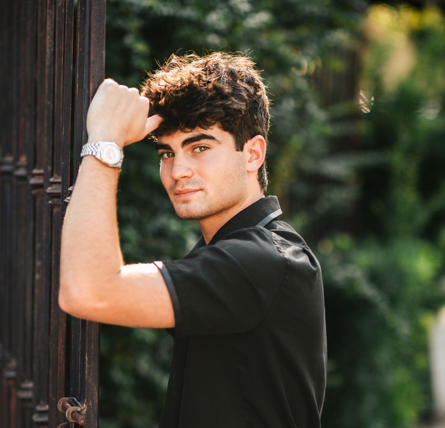 Stylish young man in black shirt leaning against iron gate wearing a silver watch.