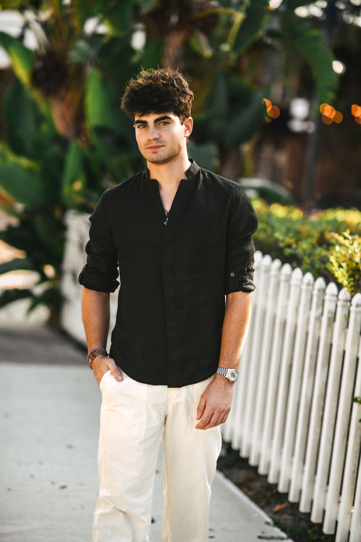 Stylish young man in black shirt and white pants walking outdoor path near white fence.