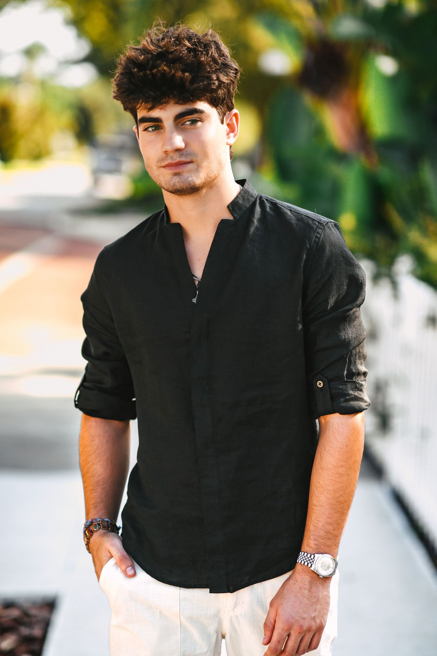 Young man in black linen shirt and white pants posing outdoors near tropical palm trees.