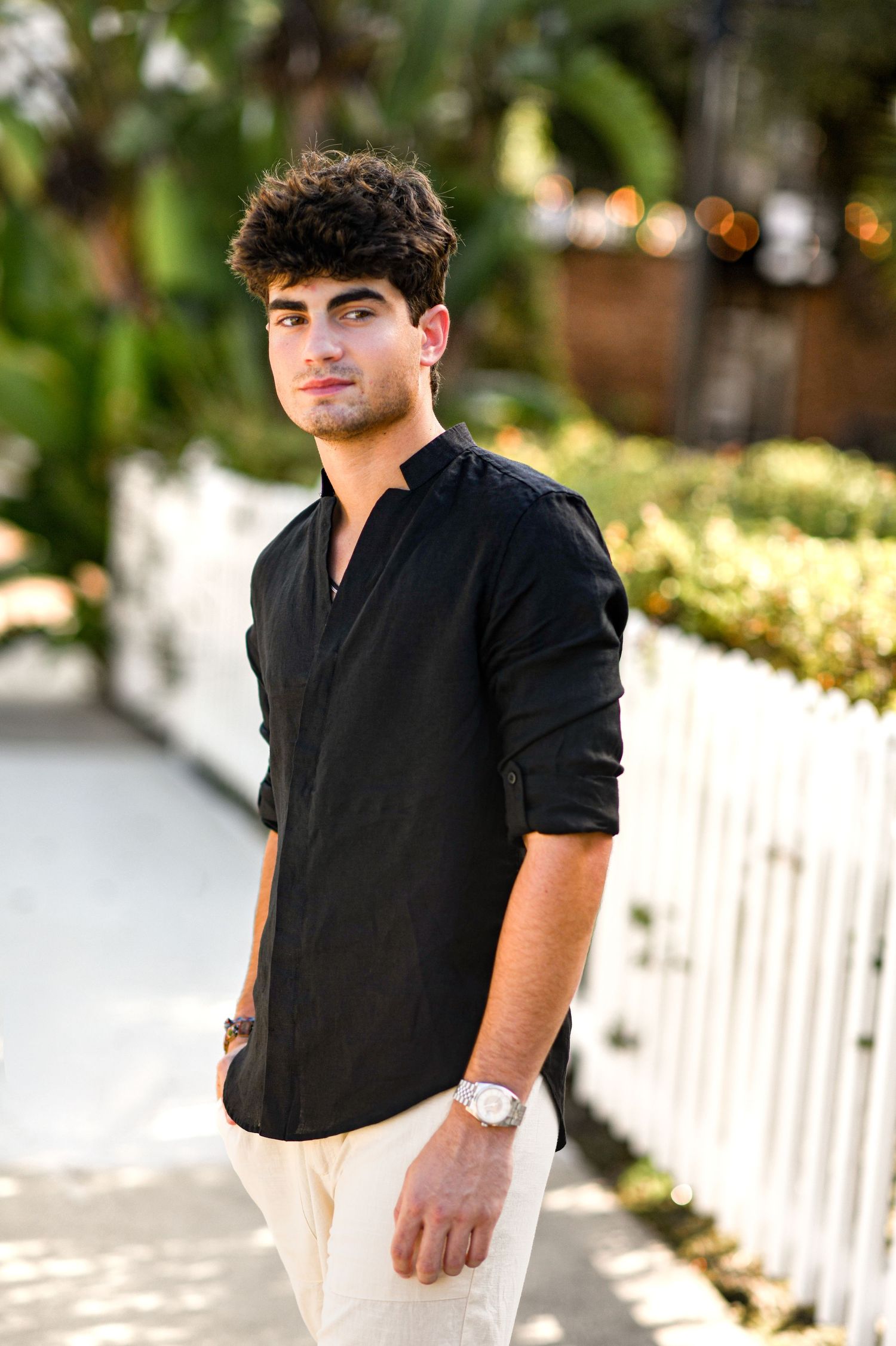Handsome young man in black button-up shirt posing on sunny outdoor walkway with white fence.
