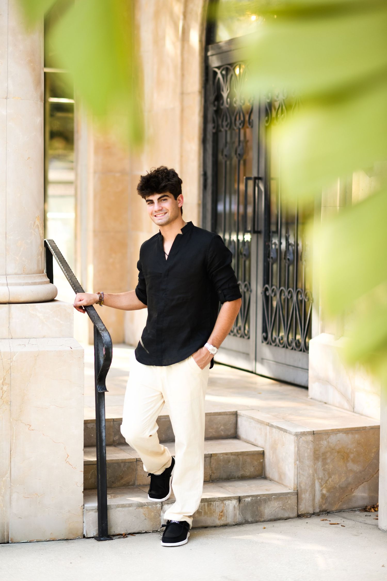 Stylish young man in black linen shirt posing on outdoor stone steps by iron railing.