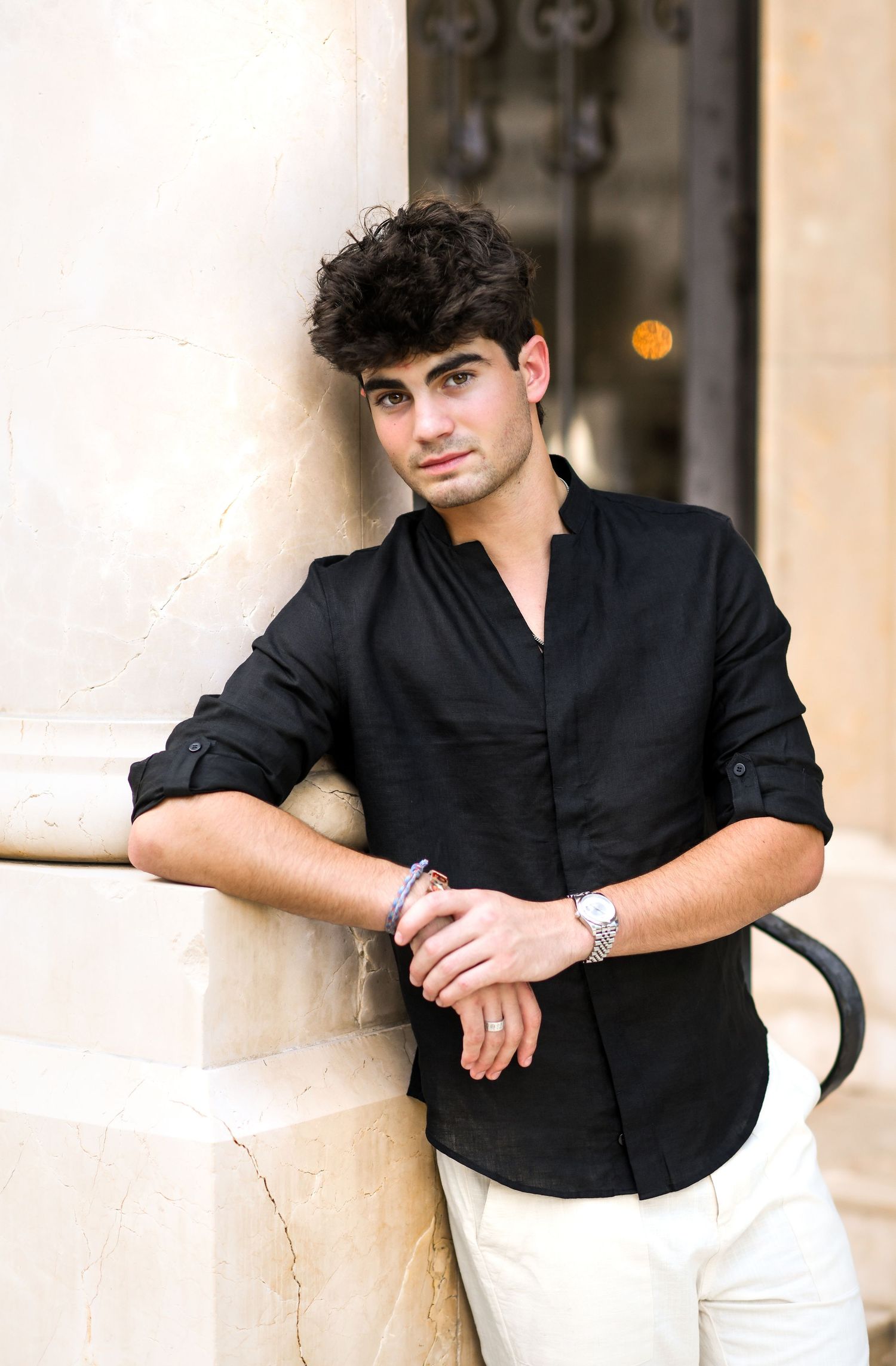 Young man in black linen shirt leaning against a stone column outdoors.