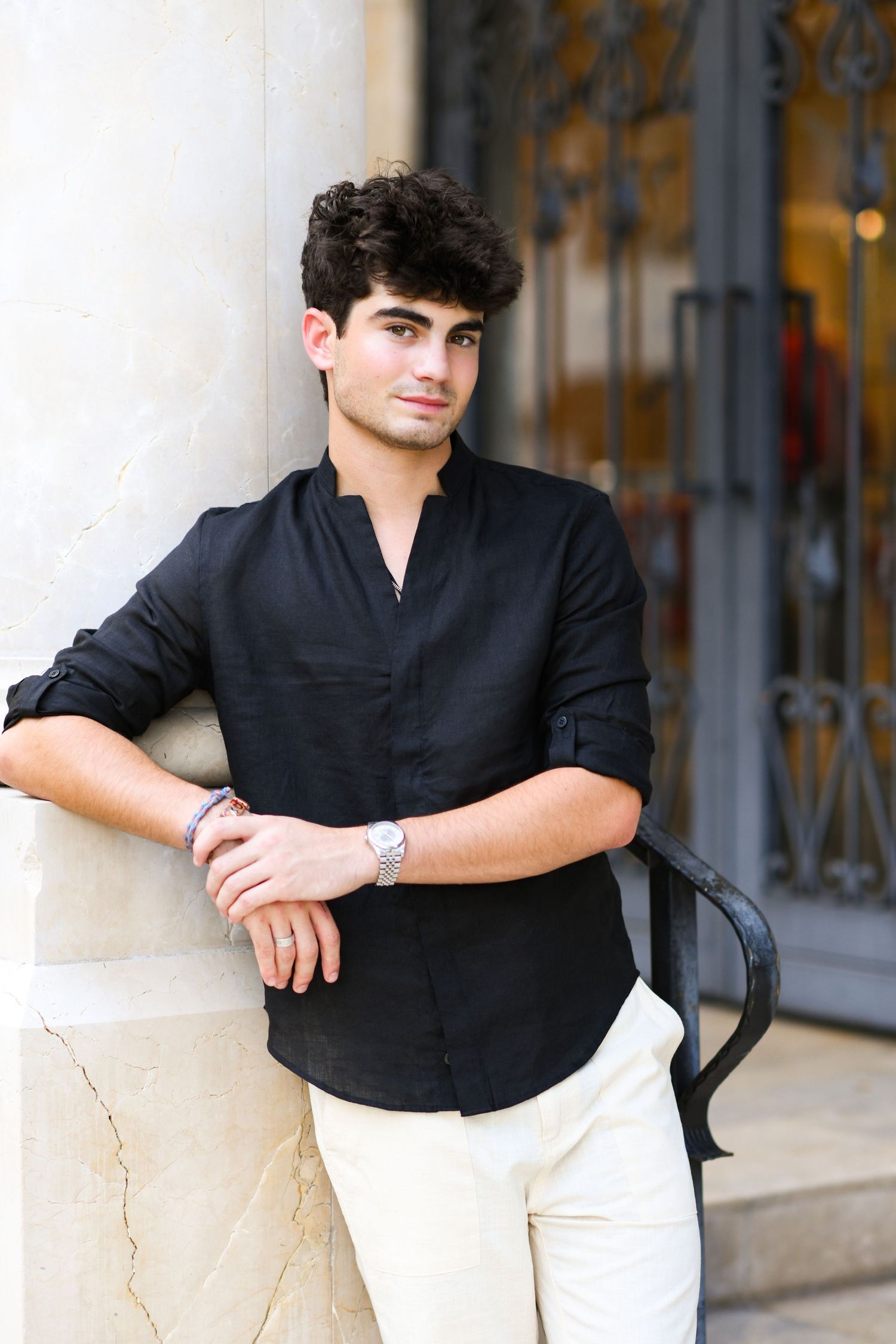 Handsome young man in black linen shirt posing against stone column outdoors.
