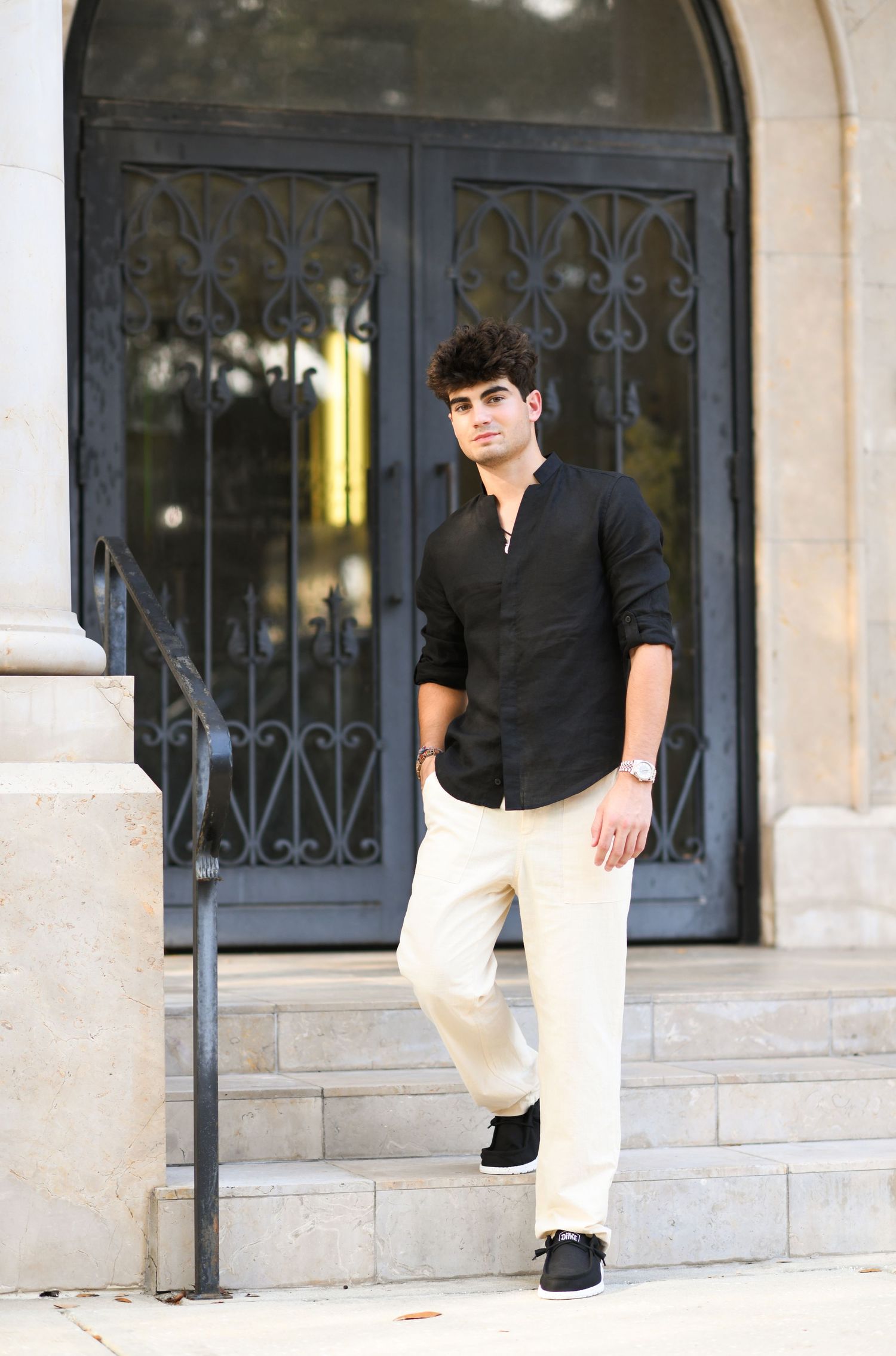 Young man in black shirt and cream pants poses on stone steps before ornate iron doors.