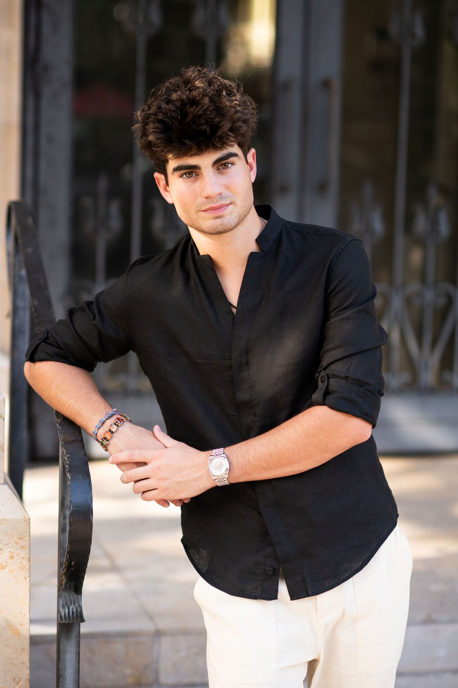 Young man in black linen shirt and white pants leaning on iron railing outdoors.