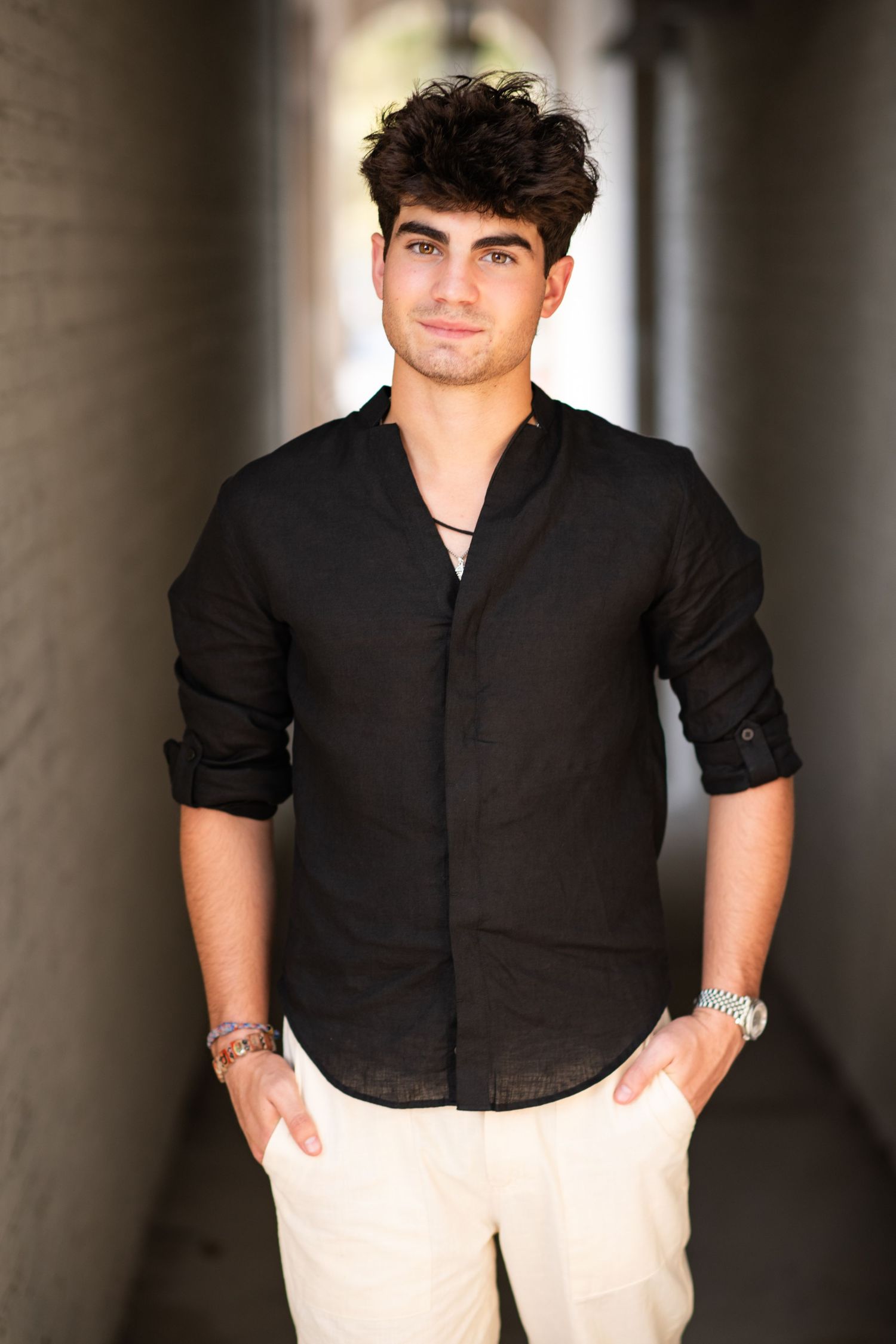 Confident young man in black linen shirt standing in outdoor corridor with hands in pockets.