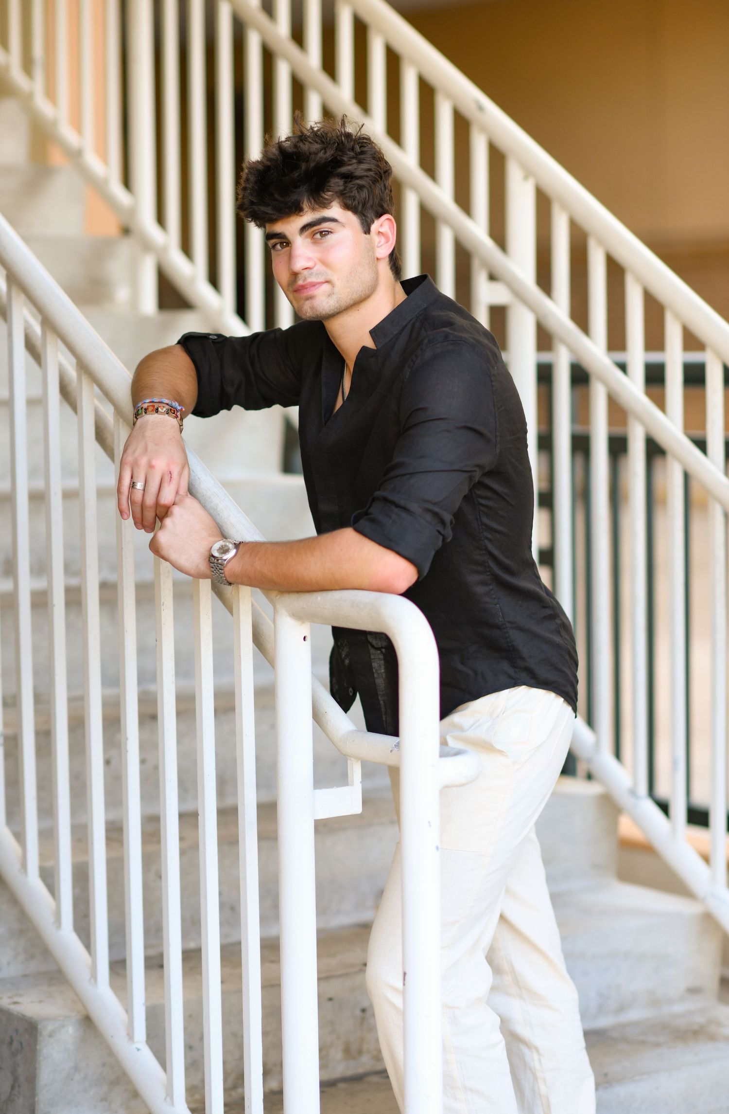 Young man in black shirt leaning on white staircase railing in a relaxed outdoor pose.