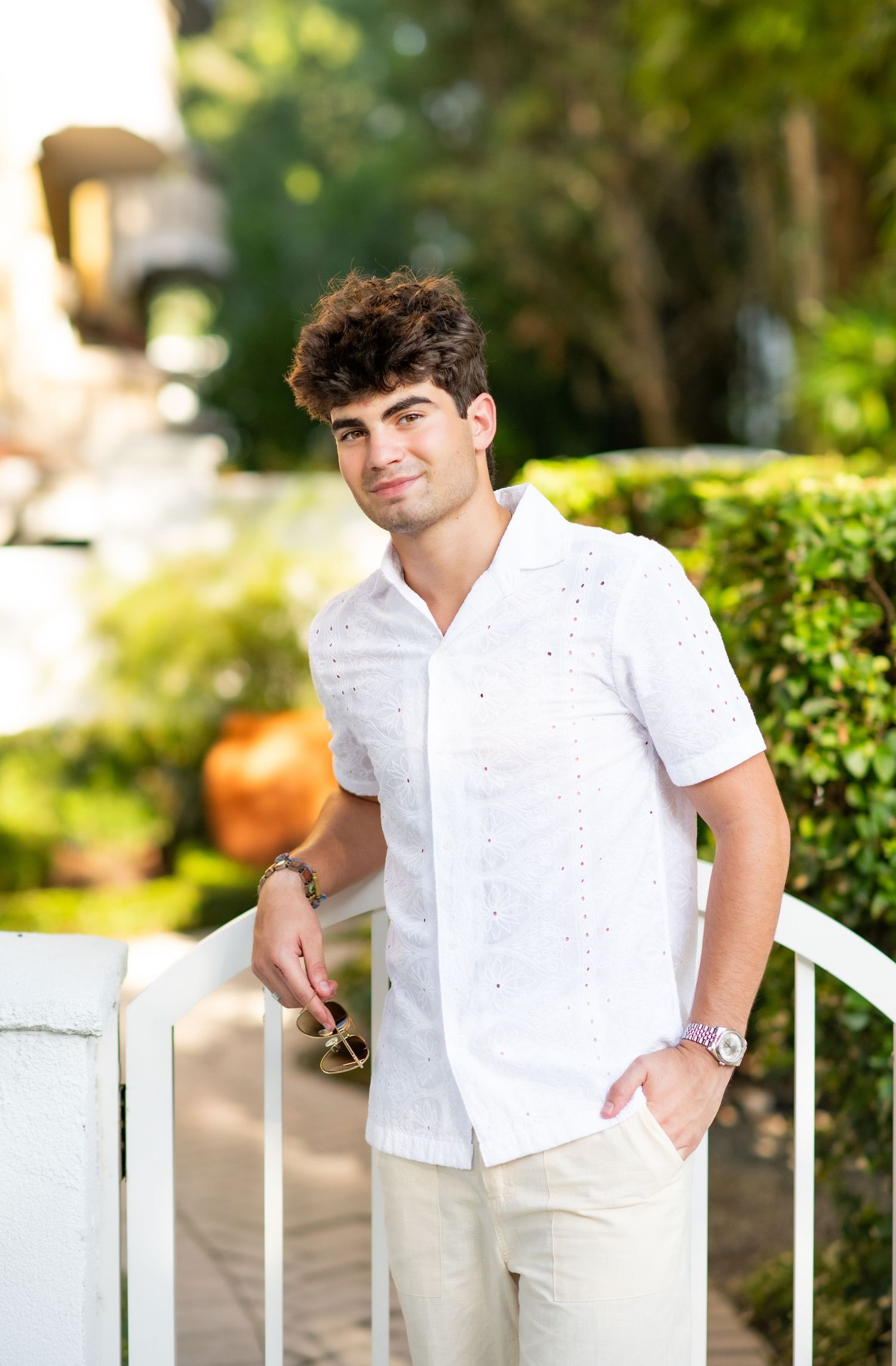 Young man in white eyelet shirt holding sunglasses by a garden fence.