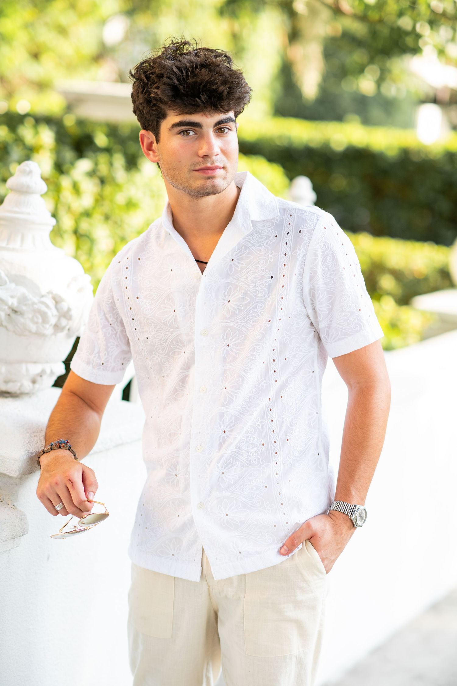 Young man in white eyelet short-sleeve shirt and khaki pants poses outdoors near white stone railing.