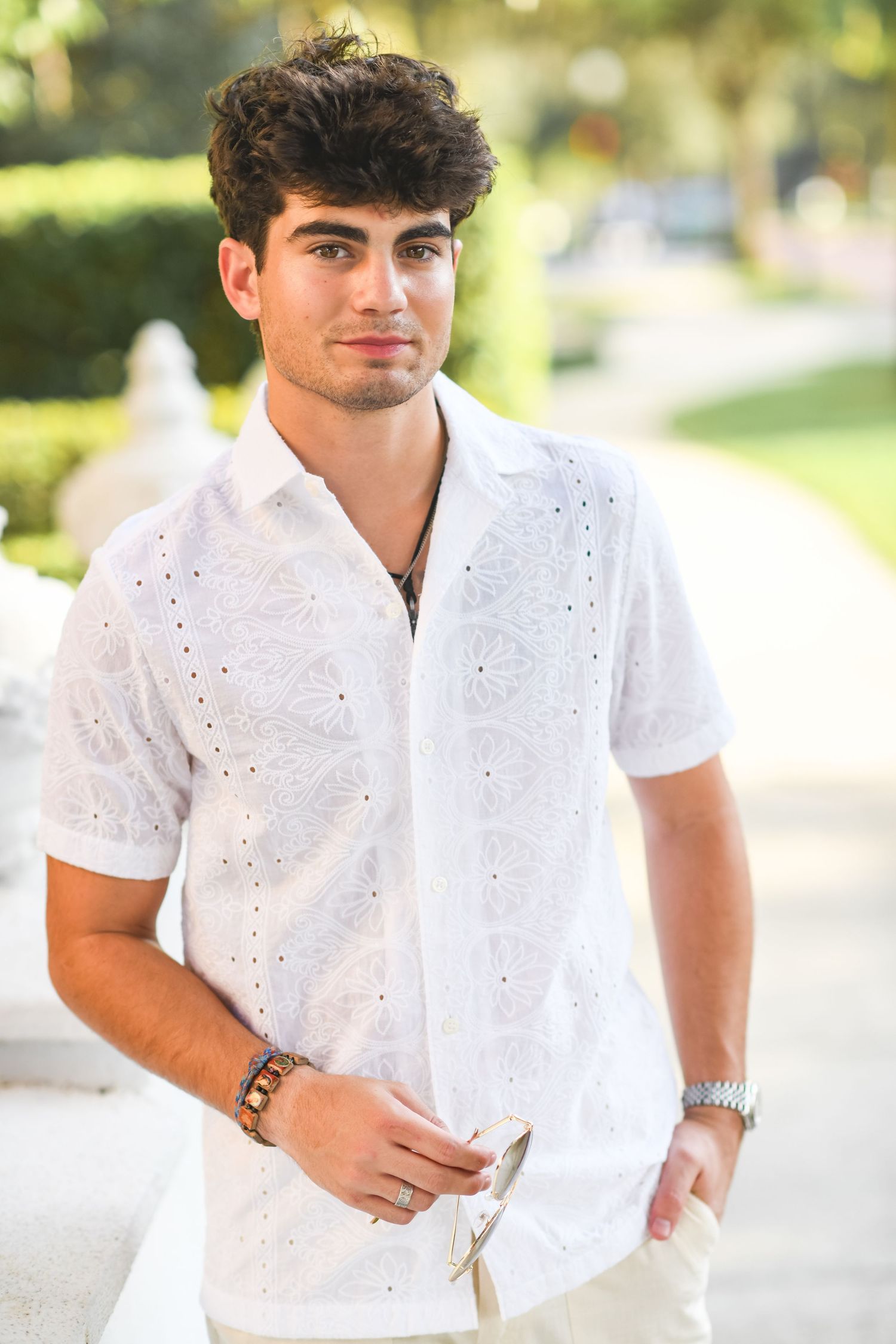 Stylish young man in white eyelet short-sleeve shirt holding sunglasses outdoors.