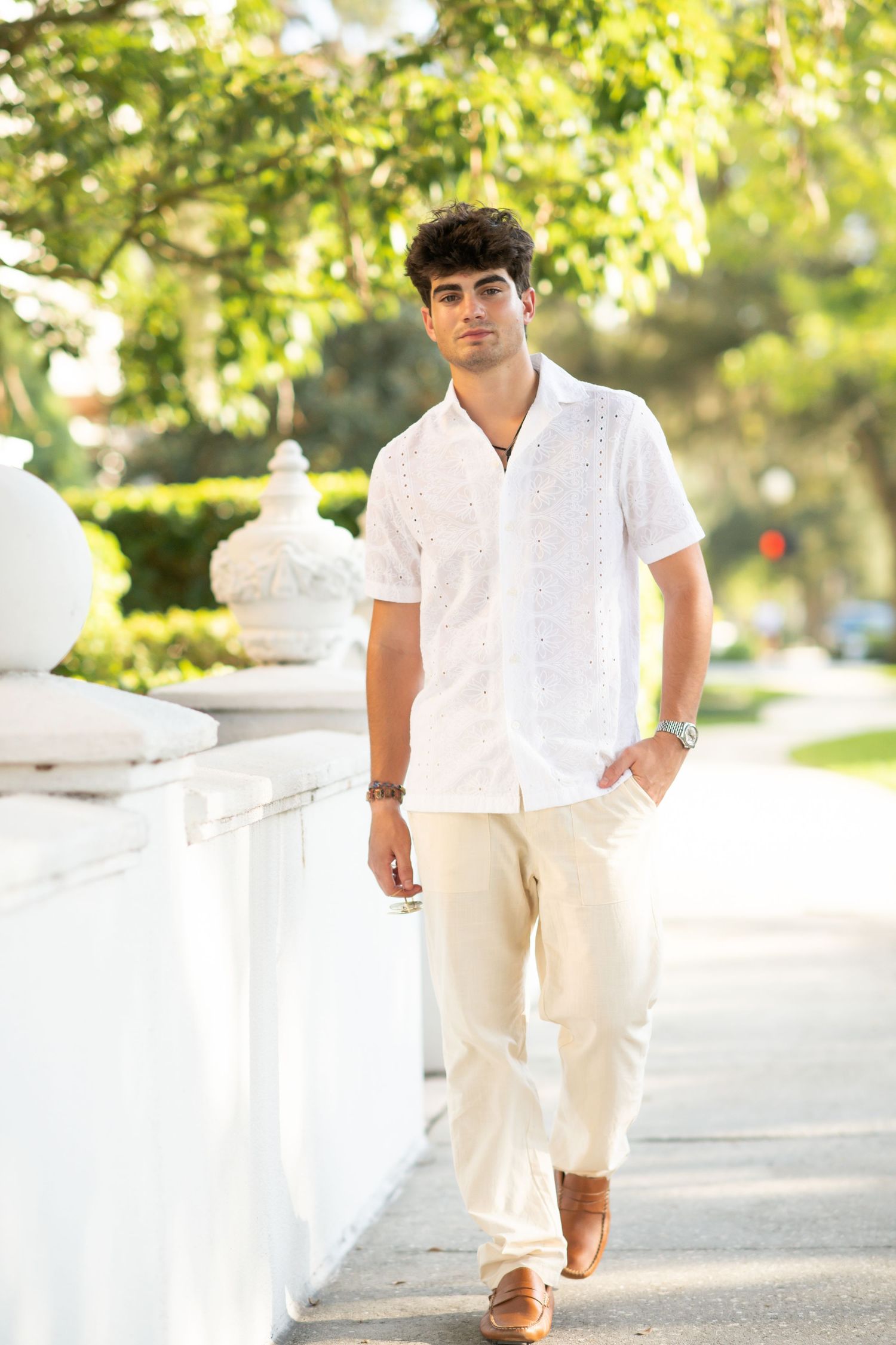 Young man in white embroidered shirt and cream pants walking along sunny outdoor pathway.