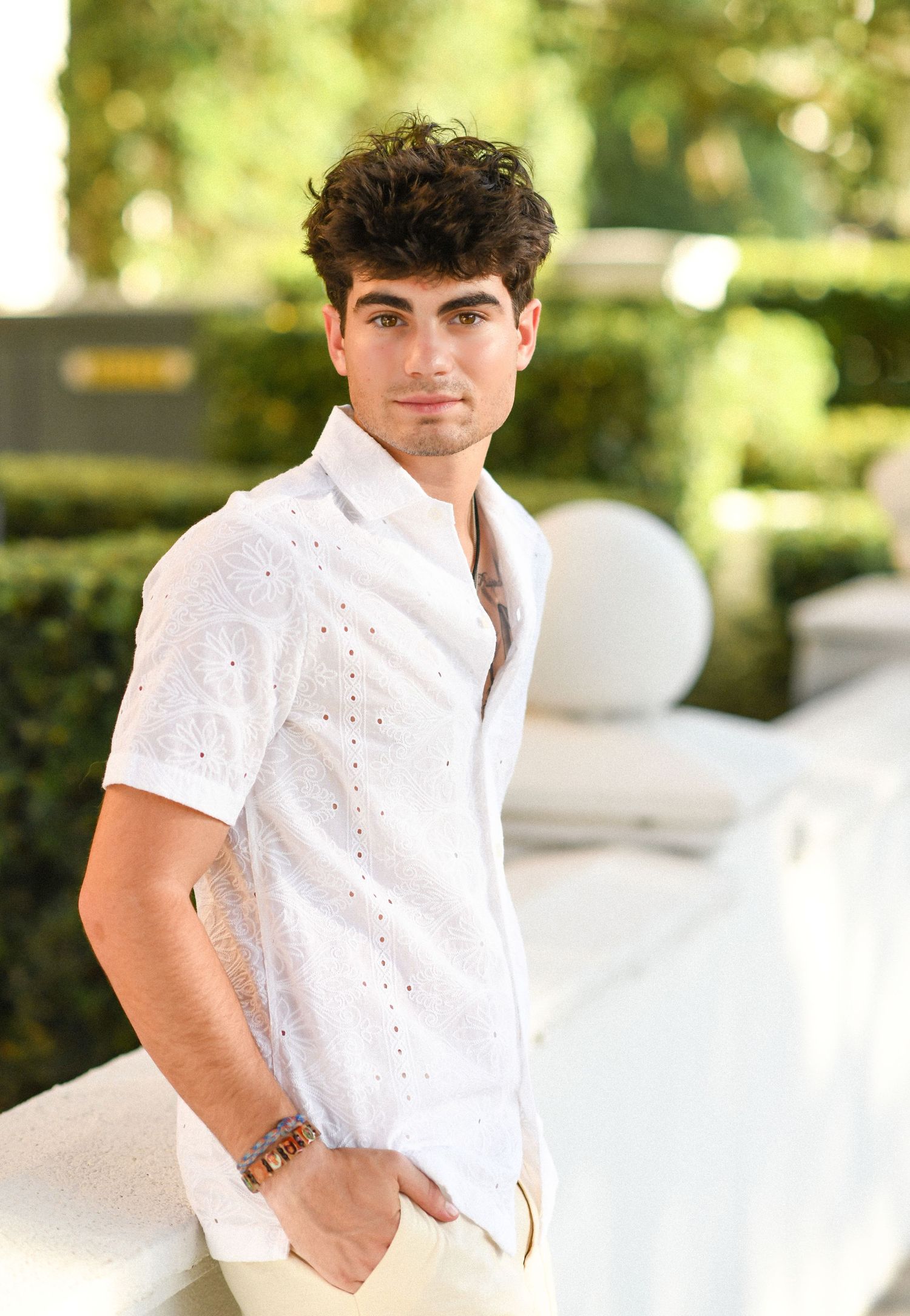 Young man in white patterned shirt leaning on railing with green hedges behind.