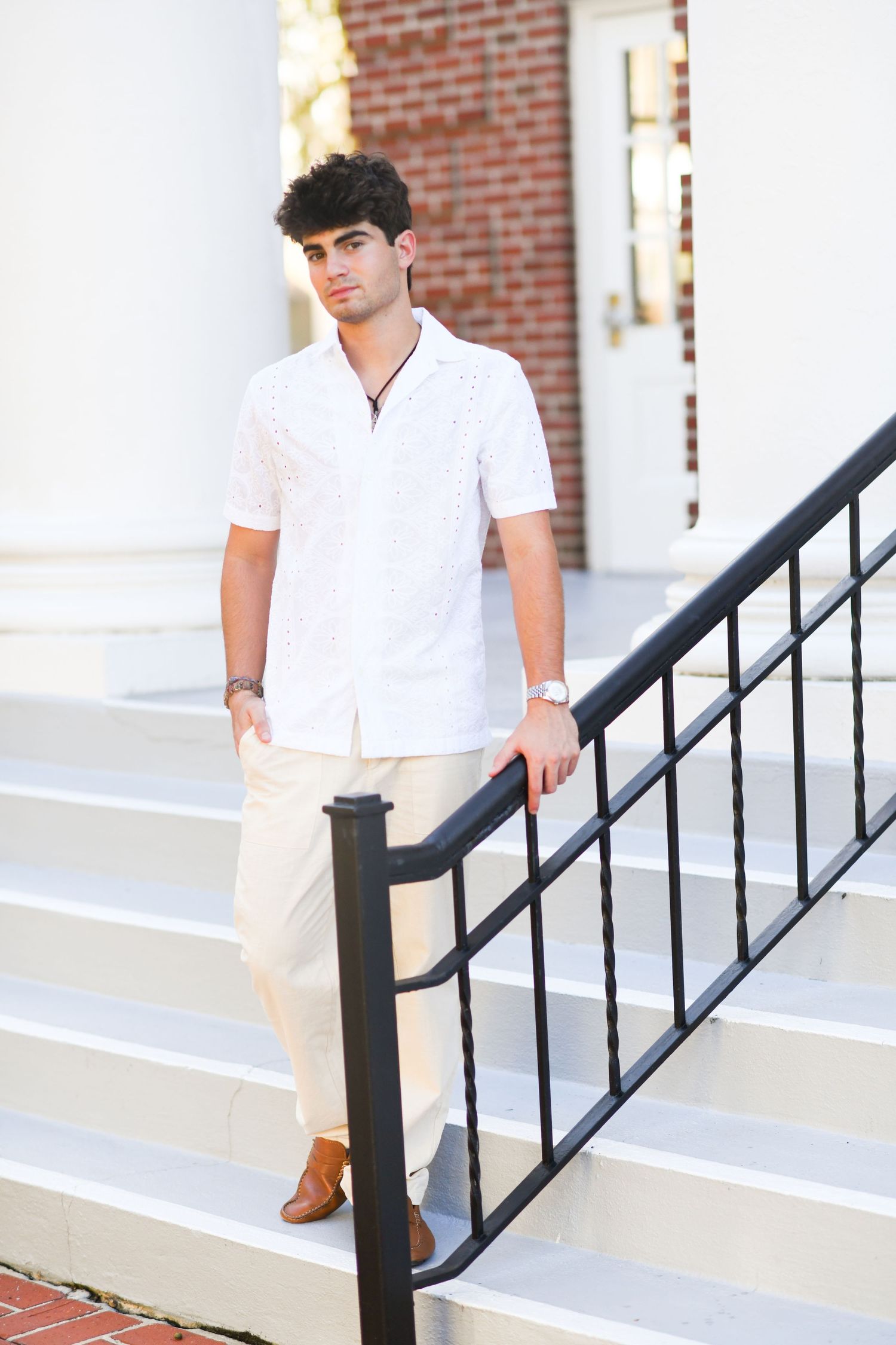 Young man in white shirt and cream pants stands on white steps holding a black railing.