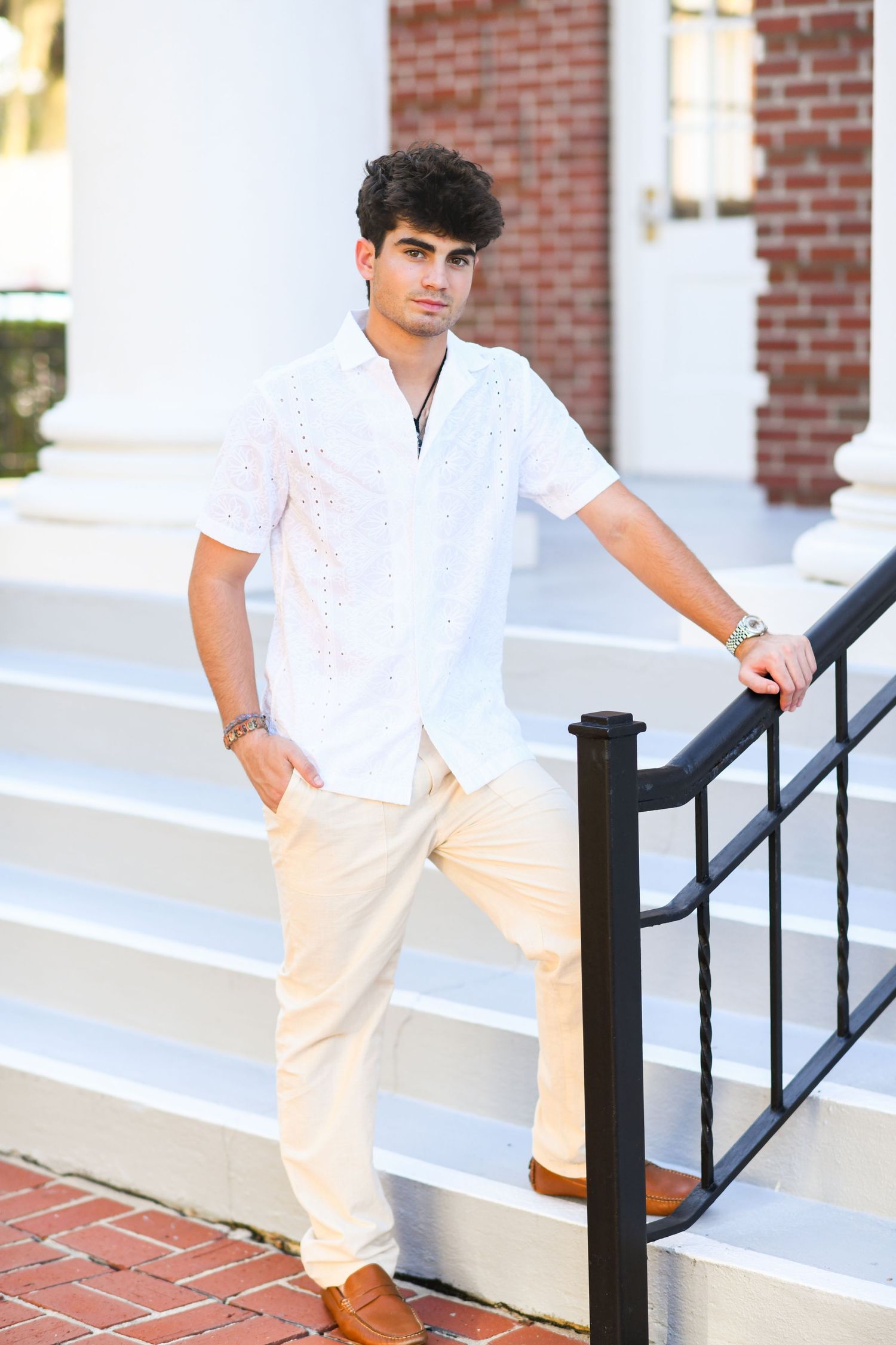 Young man in white shirt and cream pants posing on steps beside black iron railing.