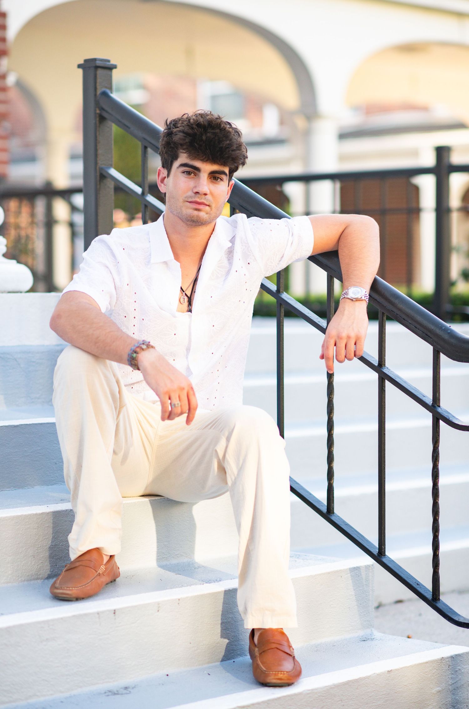 Young man in white shirt and cream pants sitting relaxed on white outdoor staircase.