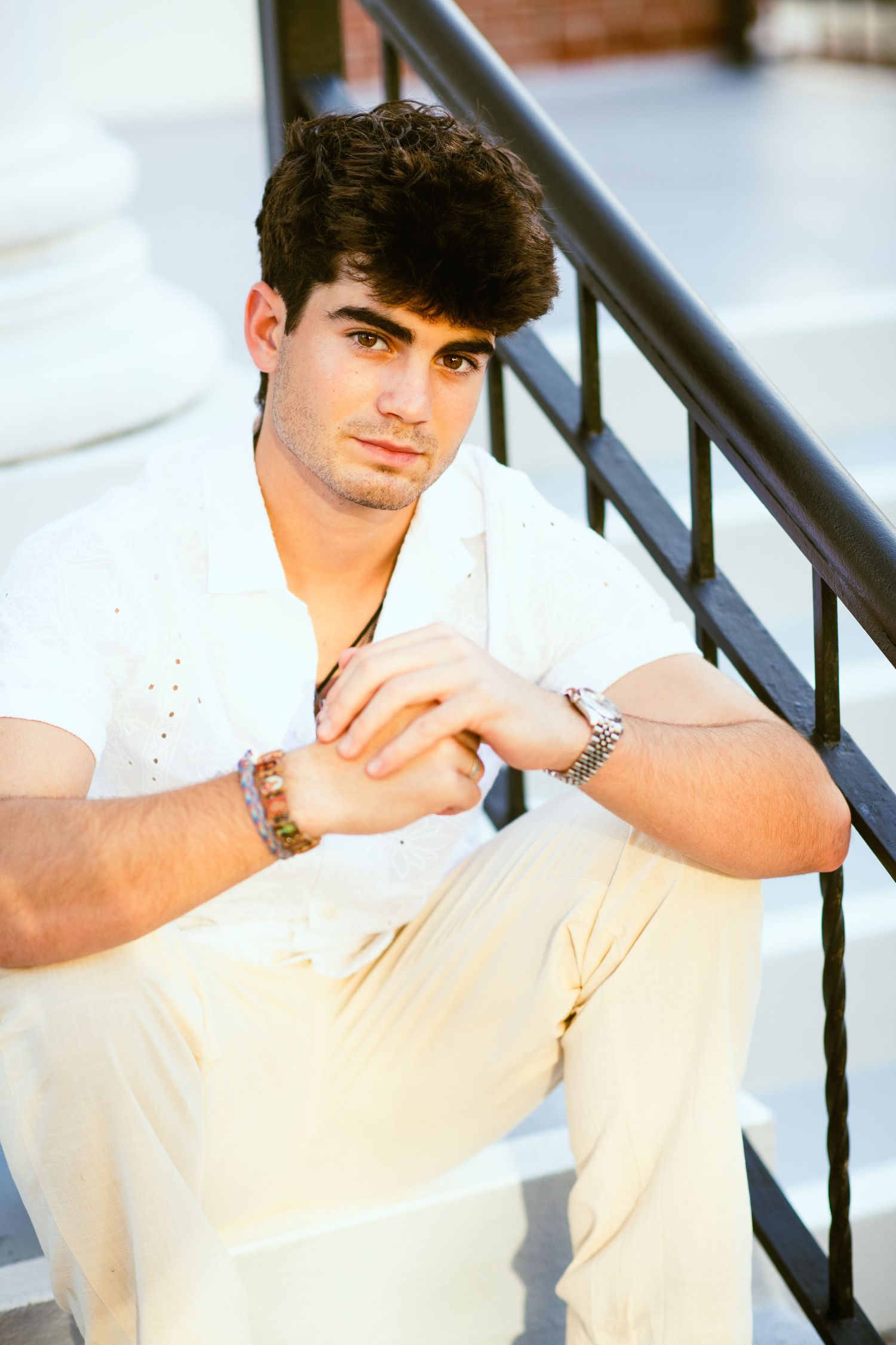 Young man in white shirt and cream pants sitting on steps with hands clasped, wearing bracelets.