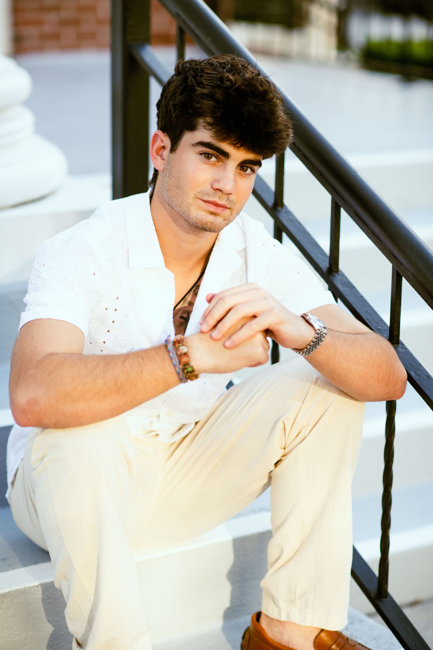 Young man in white shirt sitting on steps with hands clasped in natural light.
