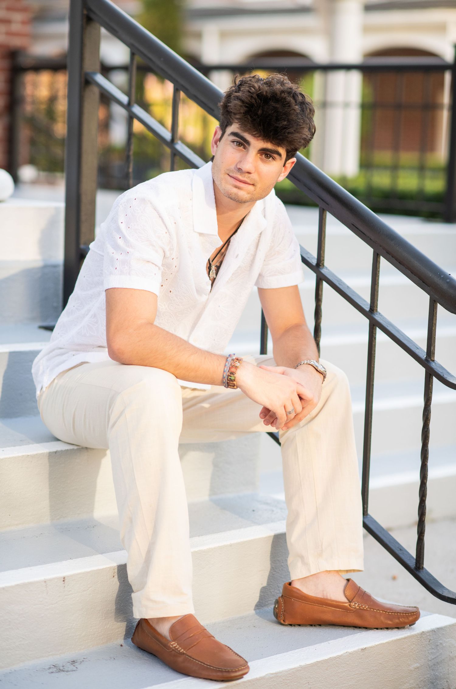 Young man in white shirt and cream pants sitting relaxed on white steps with loafers.