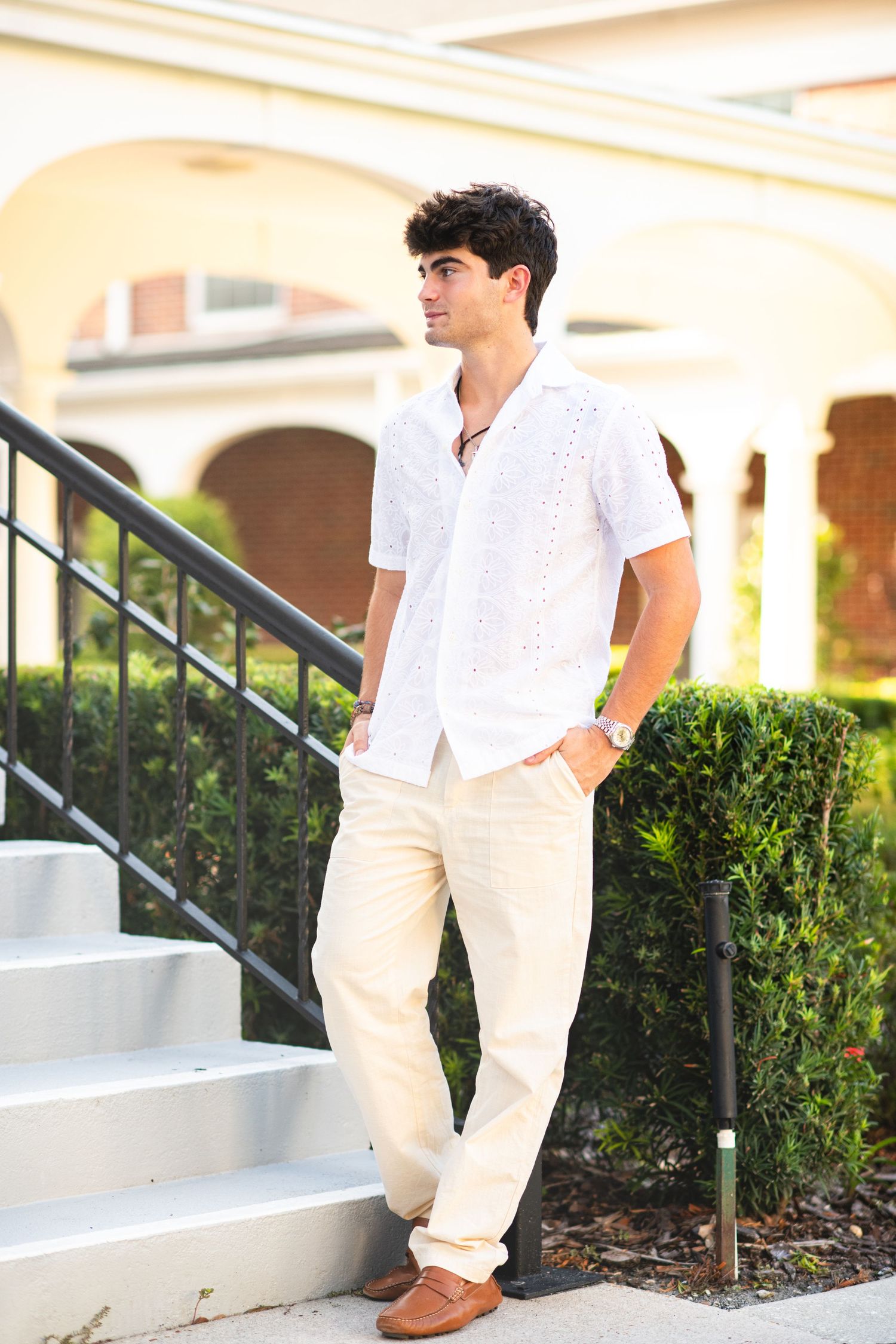 Young man in white short-sleeve shirt and cream pants standing by outdoor staircase in sunlight.