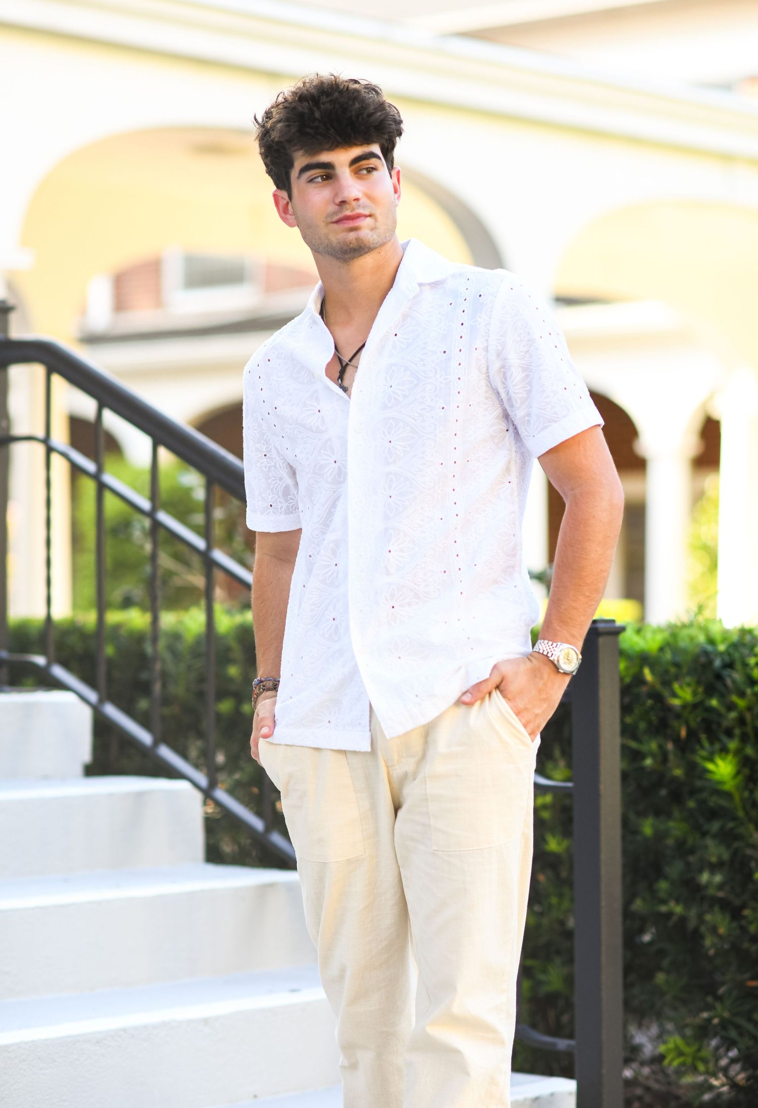 Young man in white eyelet shirt standing on outdoor steps confidently.