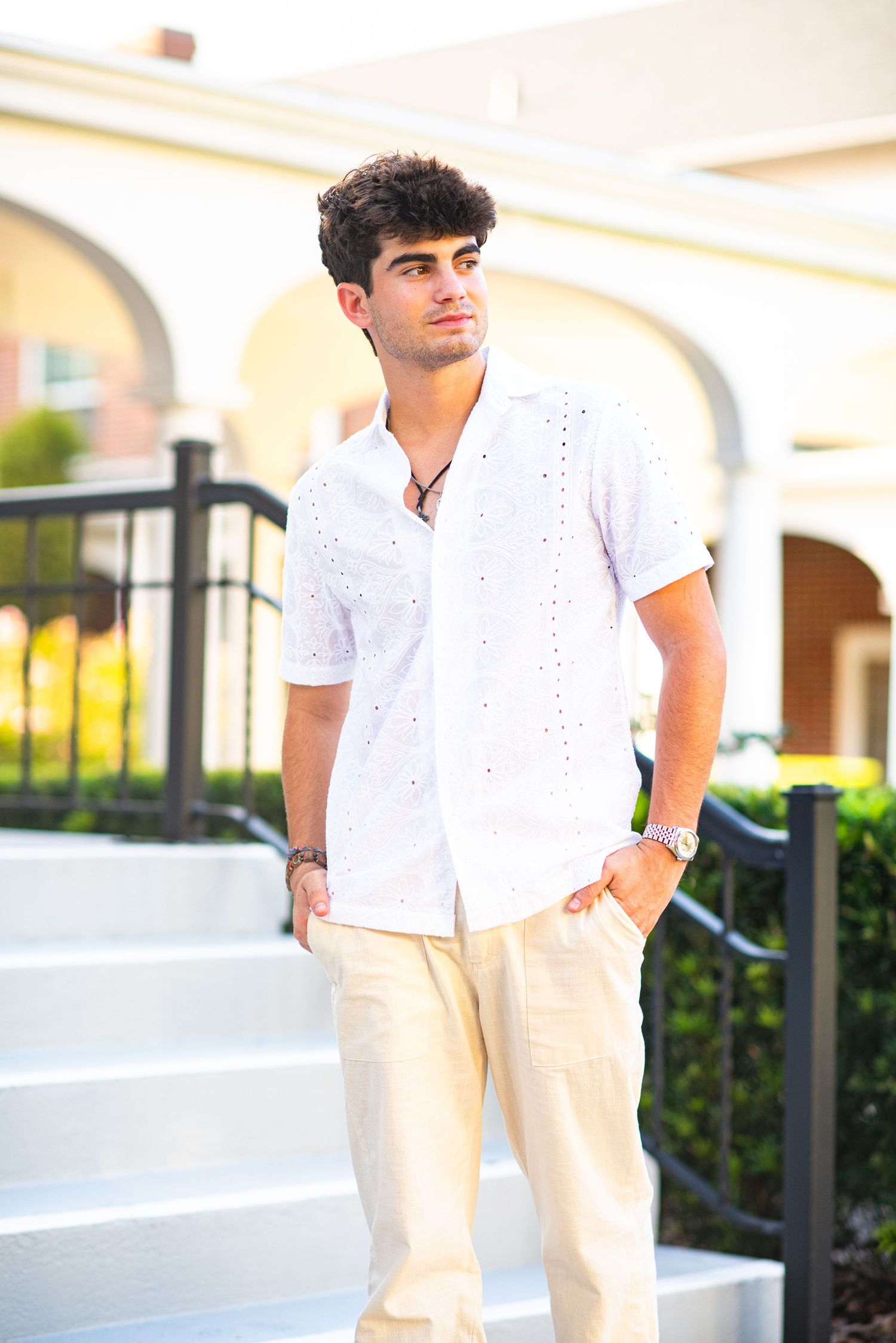 Young man in white eyelet shirt and cream pants standing outdoors near arched building.