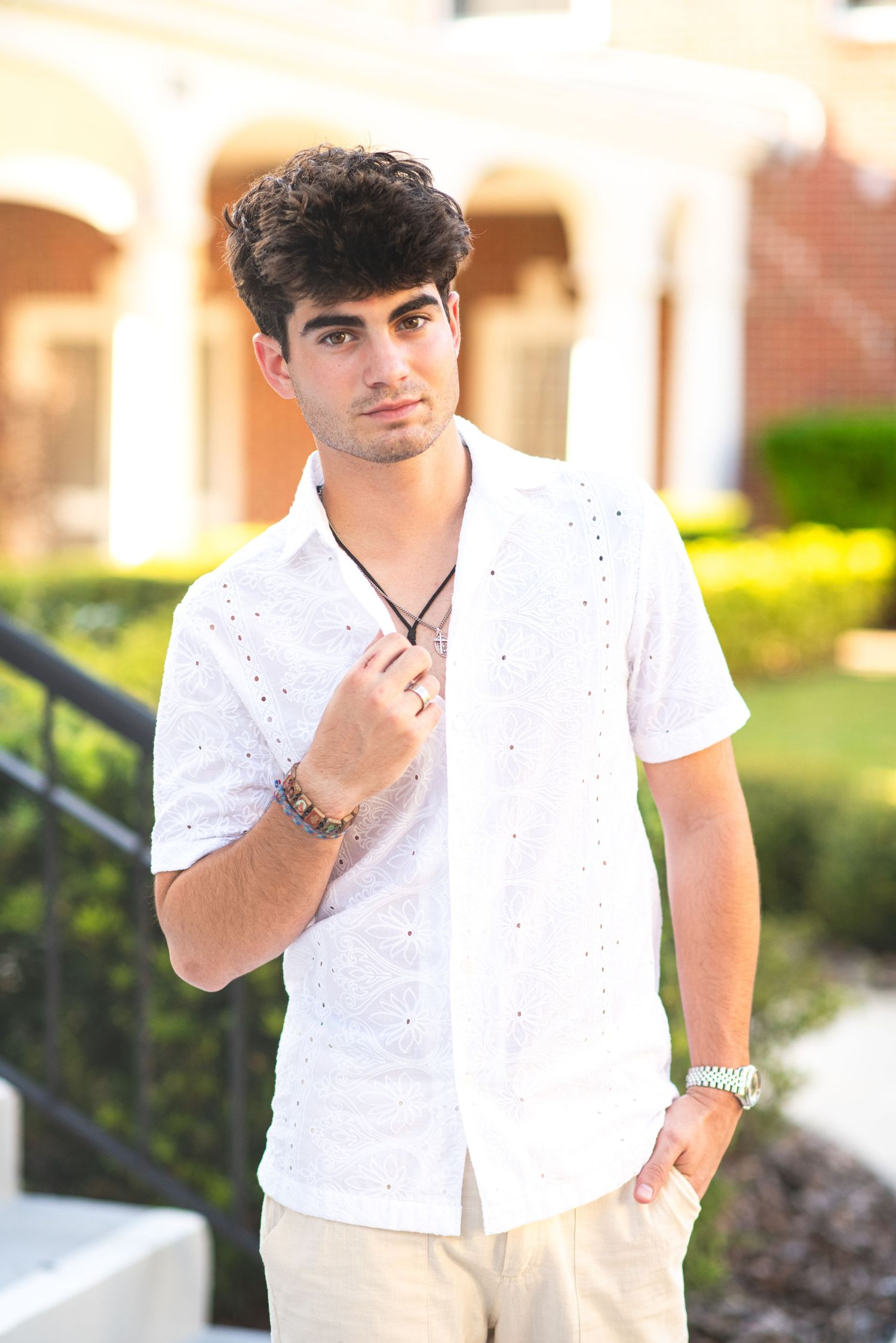Young man in white eyelet shirt holding collar while posing outdoors near building.