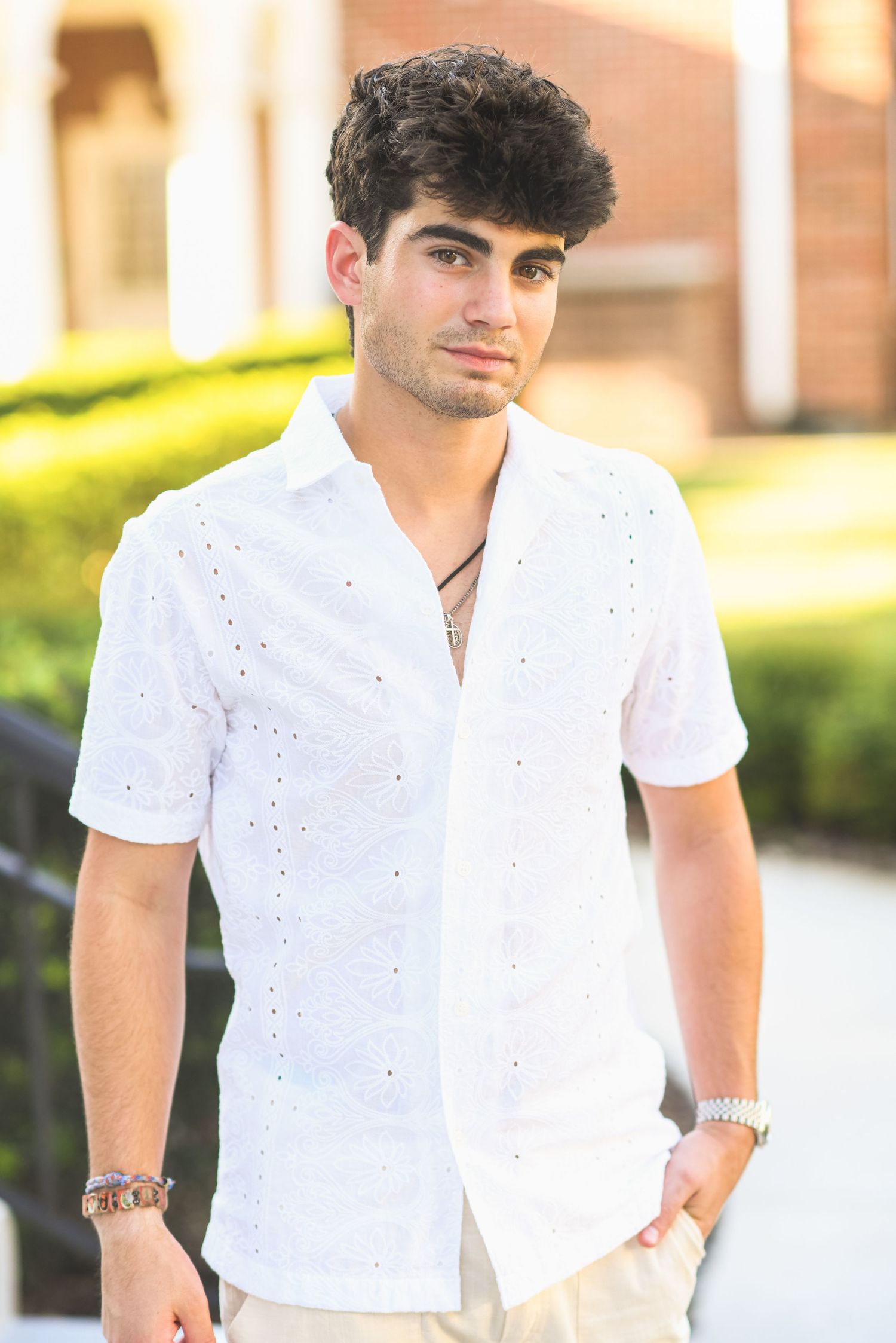Young man in white eyelet button-up shirt posing outdoors near greenery.