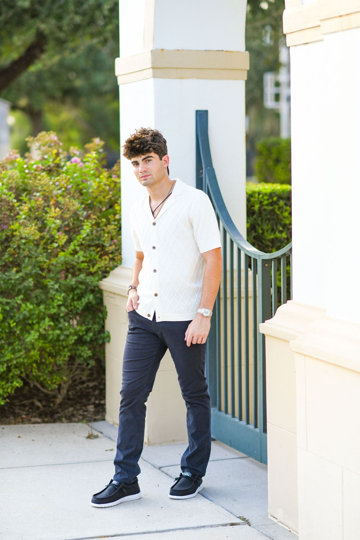 Young man in white short-sleeve shirt and dark pants standing by gate and white pillar outdoors.